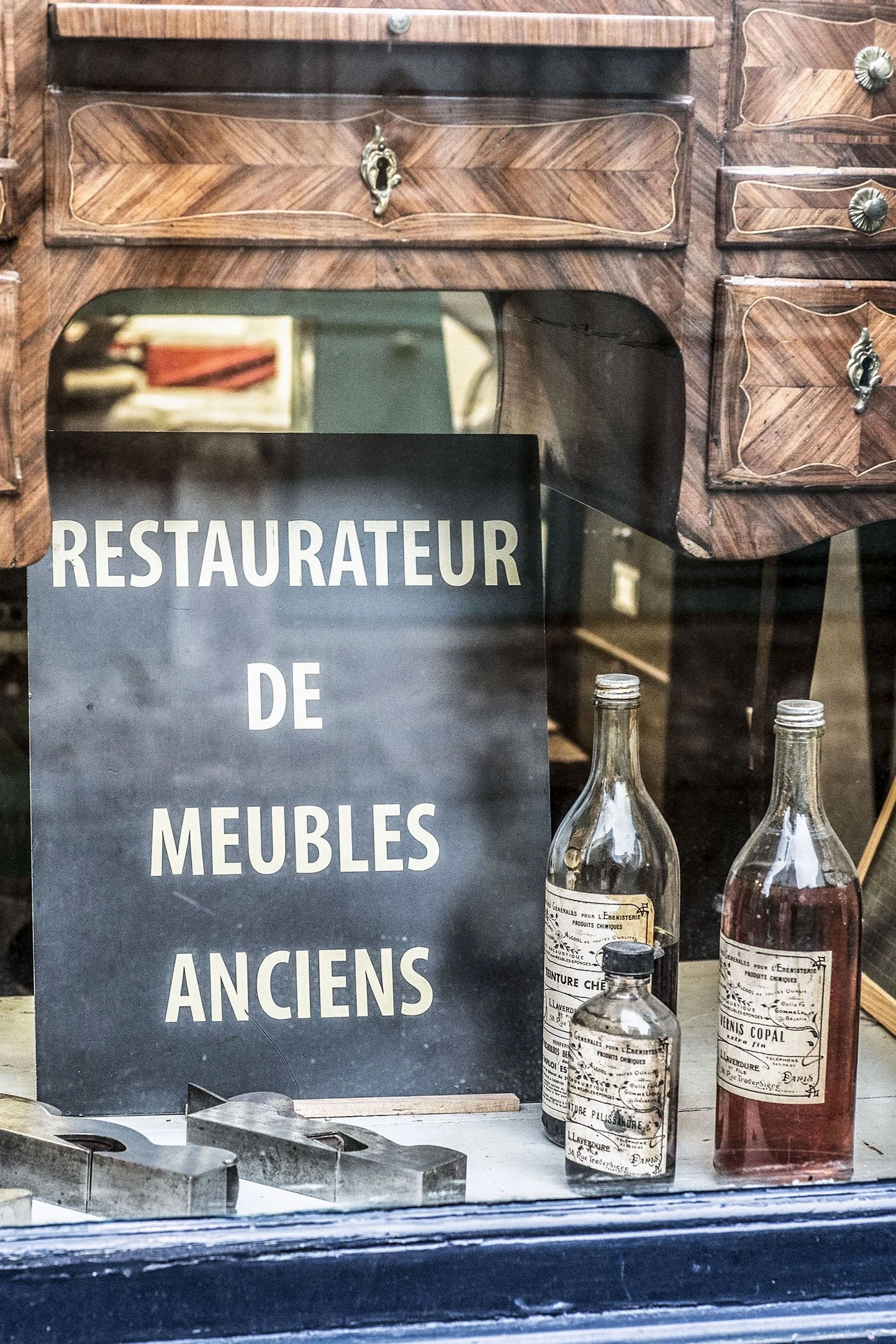 Une vitrine d'un magasin vintage avec une affiche noire indiquant 'RESTAURATEUR DE MEUBLES ANCIENS' et plusieurs bouteilles en verre avec des étiquettes anciennes posées devant l'affiche.