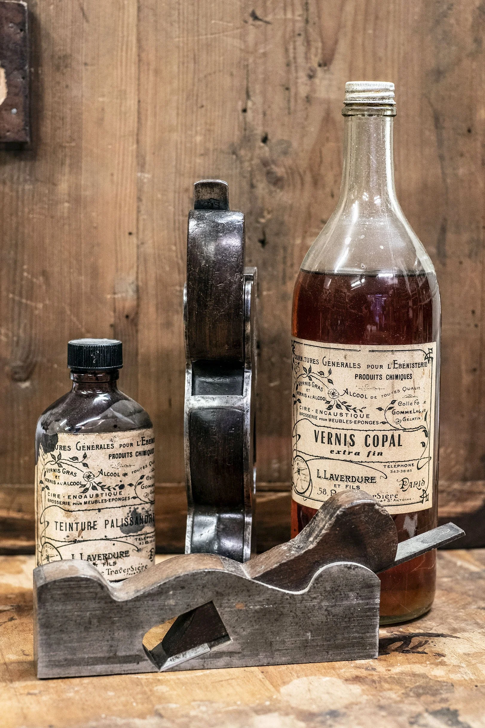 Deux bouteilles anciennes de produits chimiques et un outil en métal, posés sur une surface en bois contre un mur en bois.