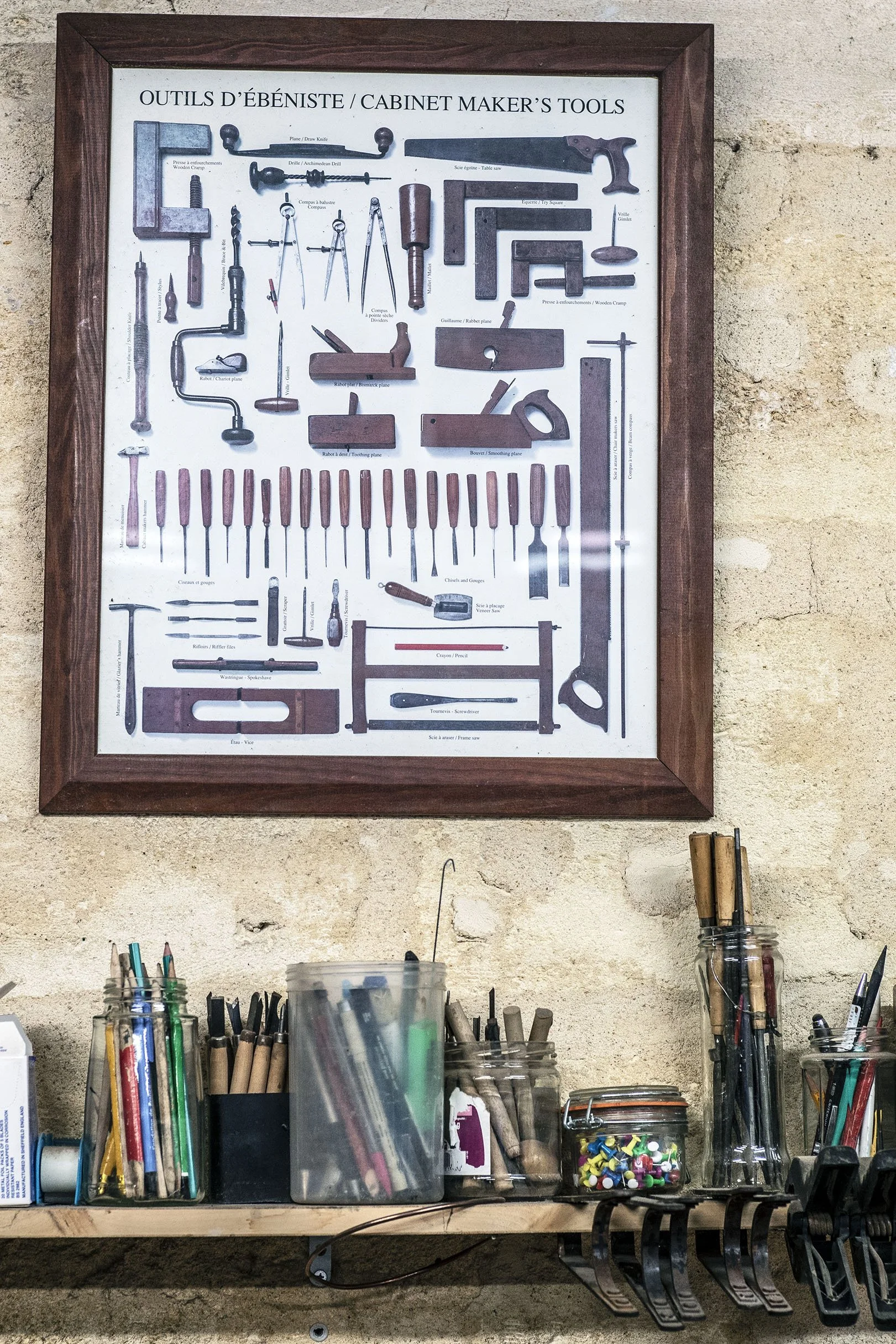 Un tableau avec différentes outils de menuiserie, comme des scies, des marteaux, des clés, des tournevis, et des outils de mesure, encadré en bois, accroché à un mur en pierre. En dessous, une étagère avec divers pinceaux, crayons, tournevis, et autr