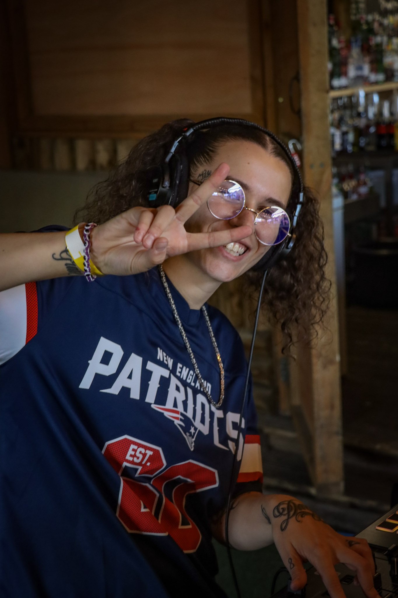 Person wearing New England Patriots shirt, round glasses, and headphones, making a peace sign and smiling at the camera, in a wooden interior with shelves in the background.