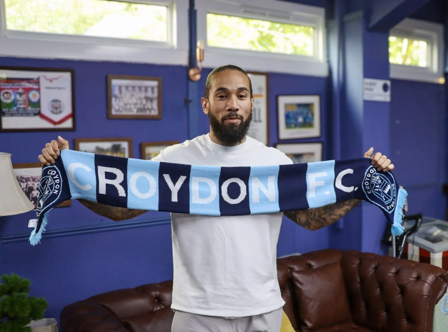 A man holding a Croydon F.C. football club scarf indoors, with blue walls and framed pictures in the background.