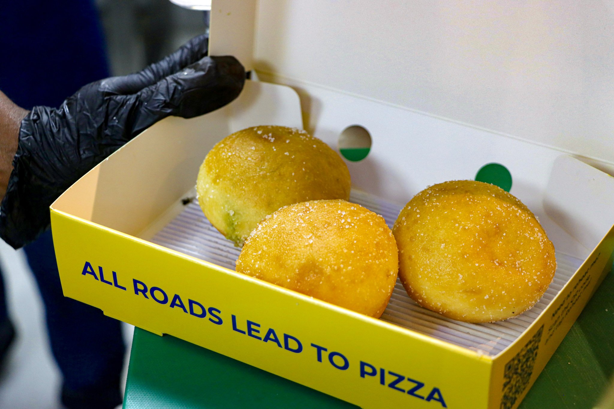 Close-up of three large, round, golden-brown fried food items in a yellow box labeled "All Roads Lead to Pizza".