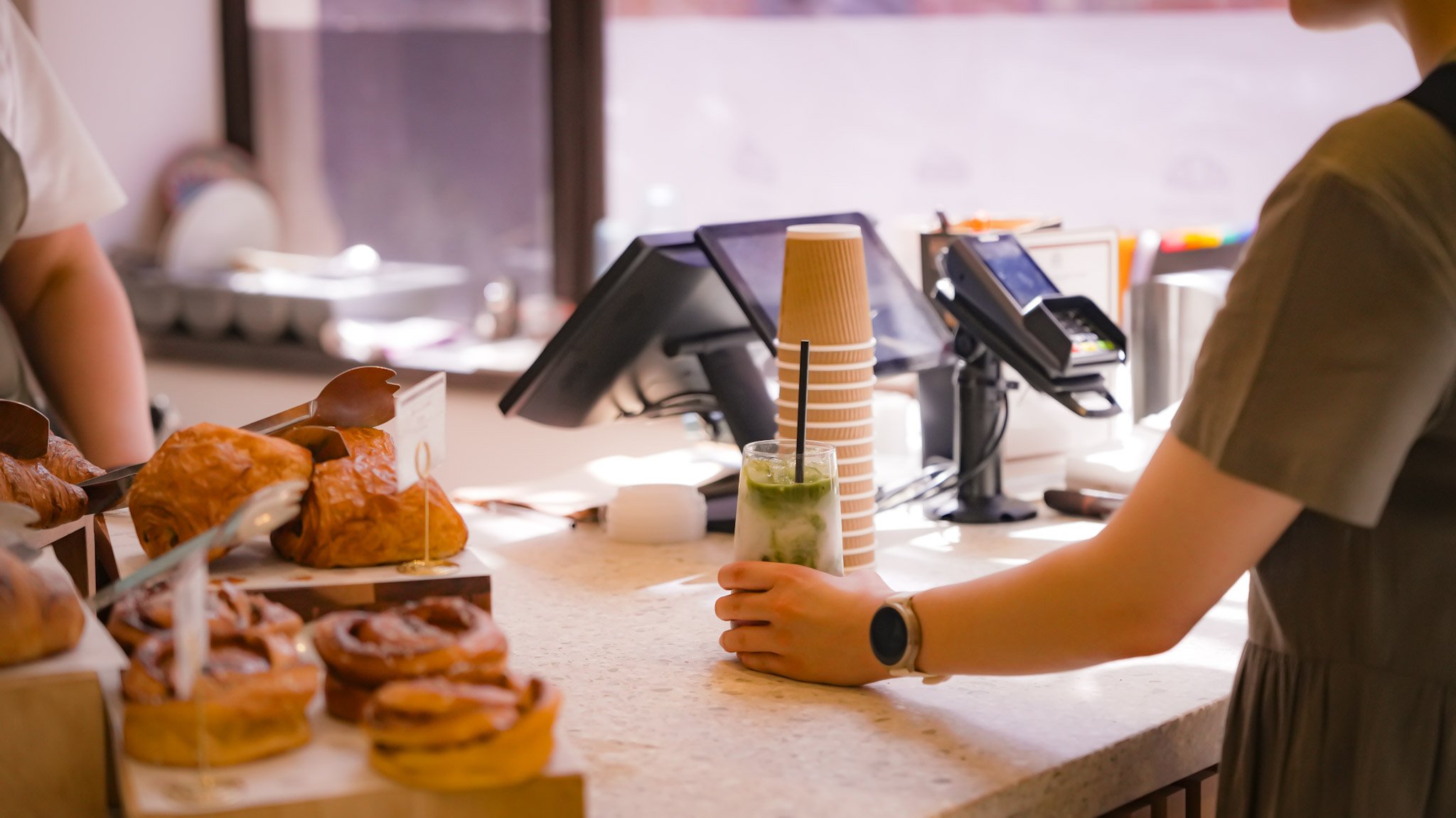 Counter at a bakery or cafe with pastries and a drink, cash register, and a person ordering.