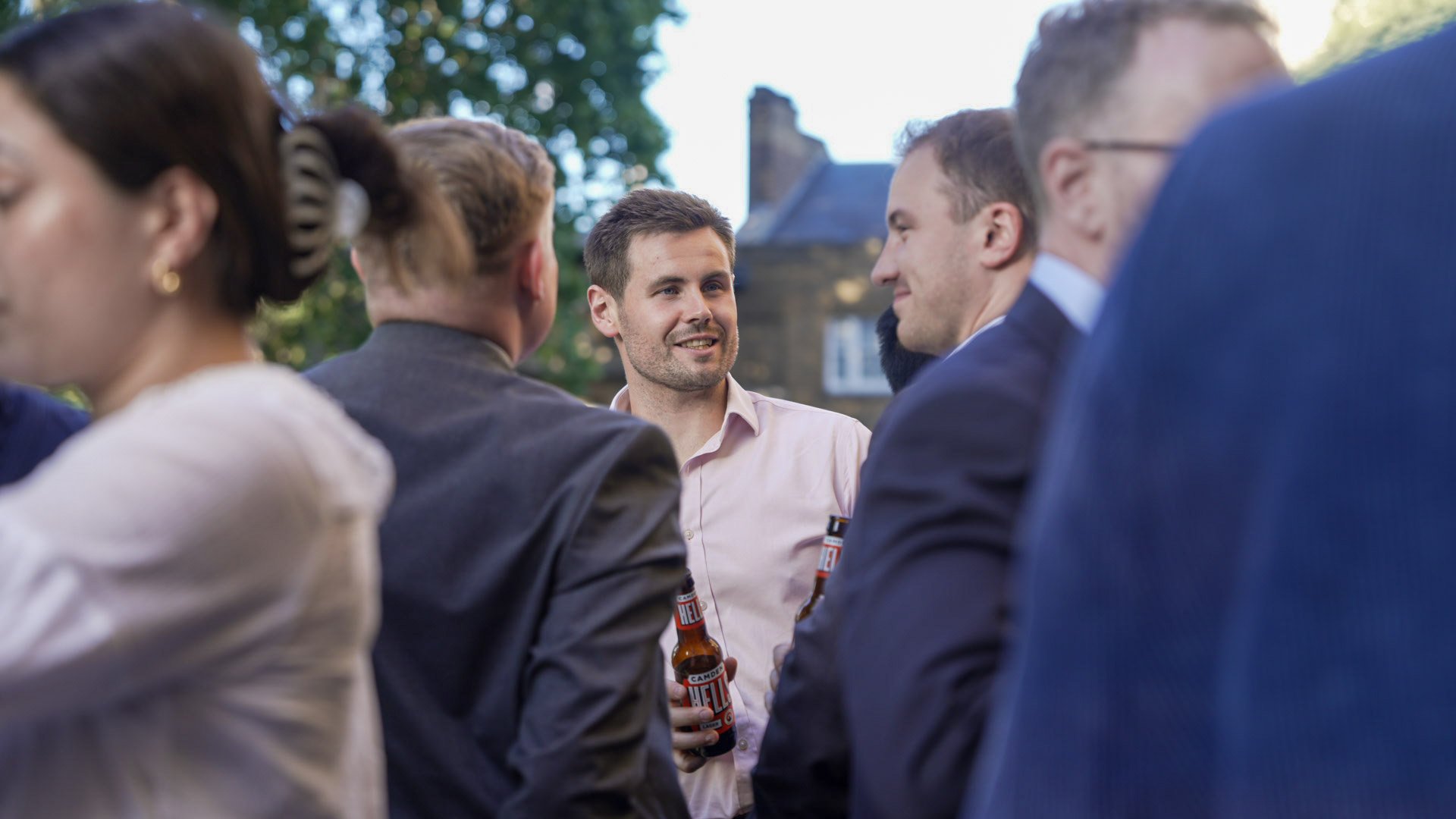 A group of people at an outdoor gathering. They are conversing and holding beer bottles. The focus is on a young man in a light pink shirt talking to others. The background includes trees and a house.