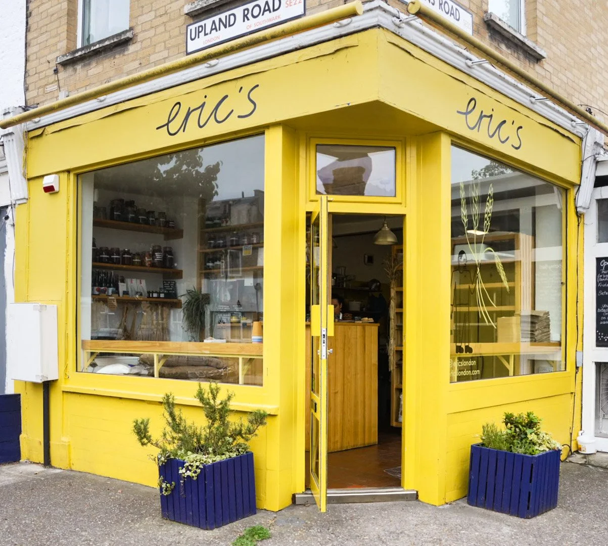 Bright yellow storefront of Eric's shop with large front windows, potted plants outside, and a sign with the shop name above the entrance. Inside, shelves with jars and a wooden counter are visible.