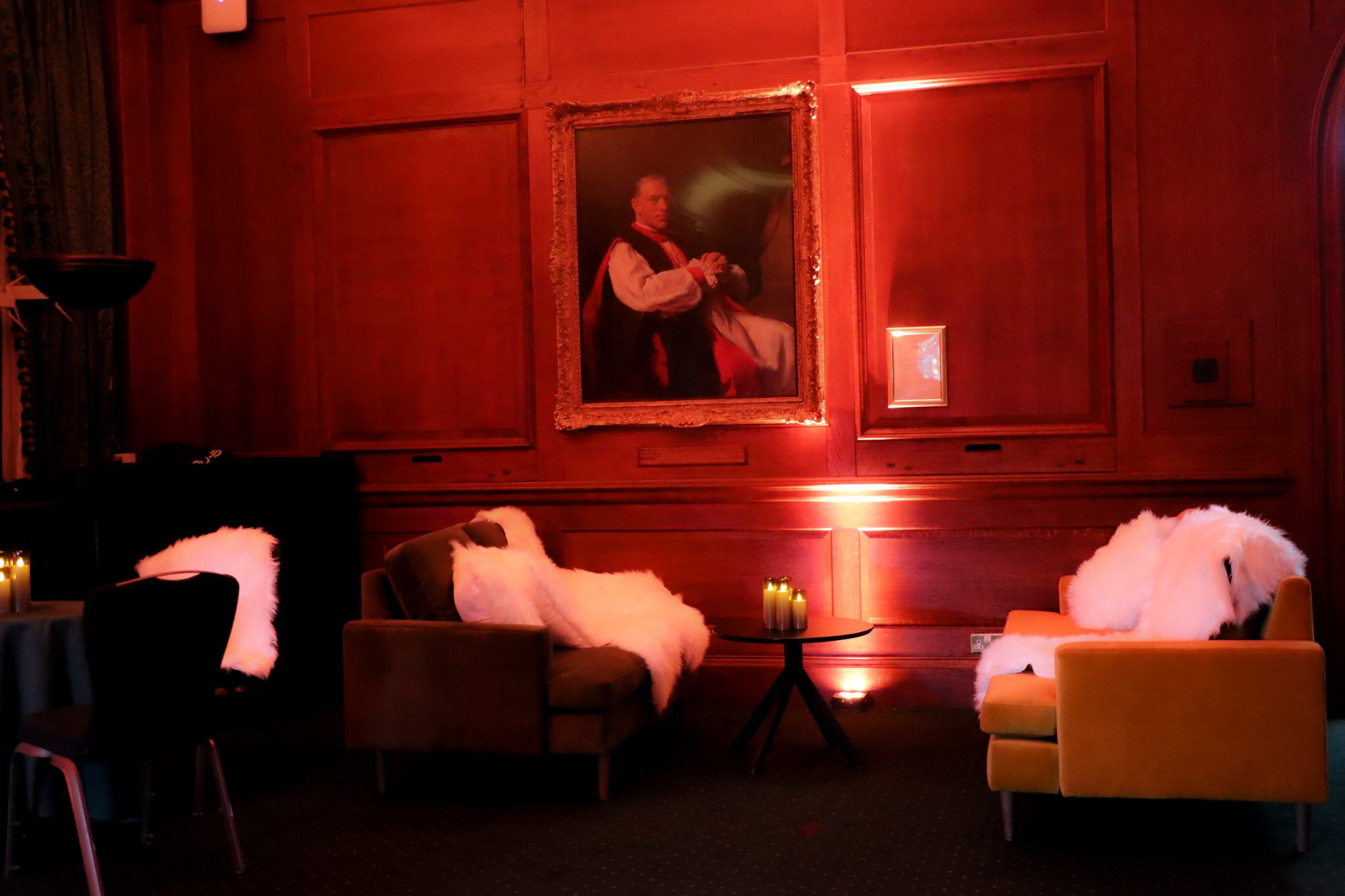 A dimly lit lounge with wooden-paneled walls, featuring a large framed portrait of a historical man dressed in 18th-century attire. The room has plush chairs with white fur throws, small round tables with candles, and a warm, cozy ambiance.