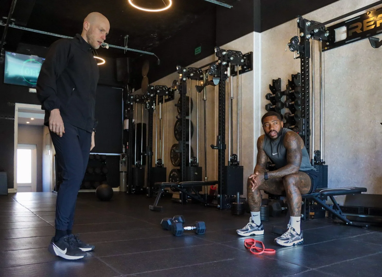 A man in workout attire sitting on a bench in a gym, with a trainer standing nearby. Gym equipment such as dumbbells, resistance bands, and weight racks are visible.