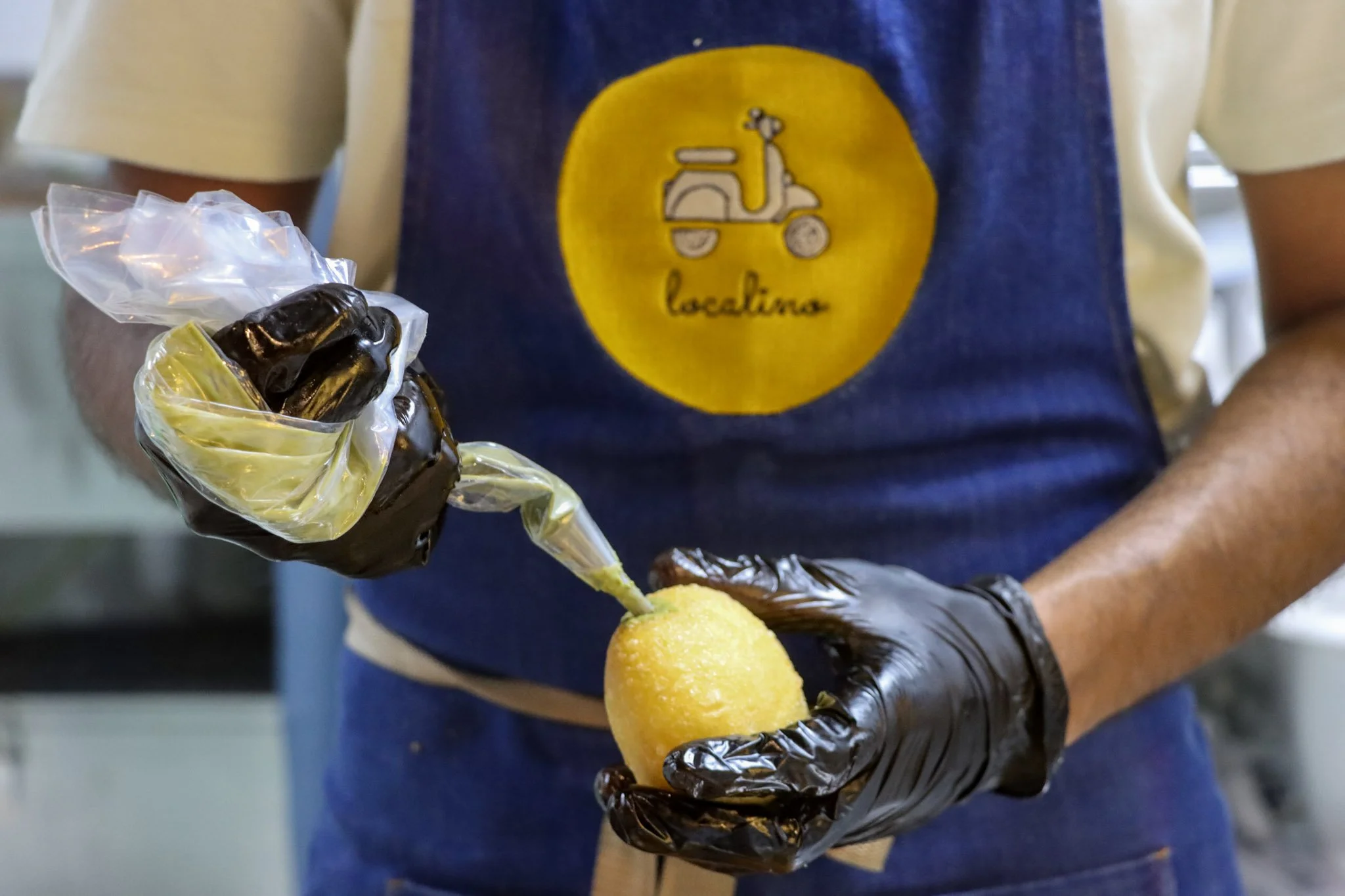 A person wearing a blue apron with a yellow circle logo that says 'localine' and black gloves is holding a yellow food item on a stick, covered with a clear plastic glove, in an indoor setting.