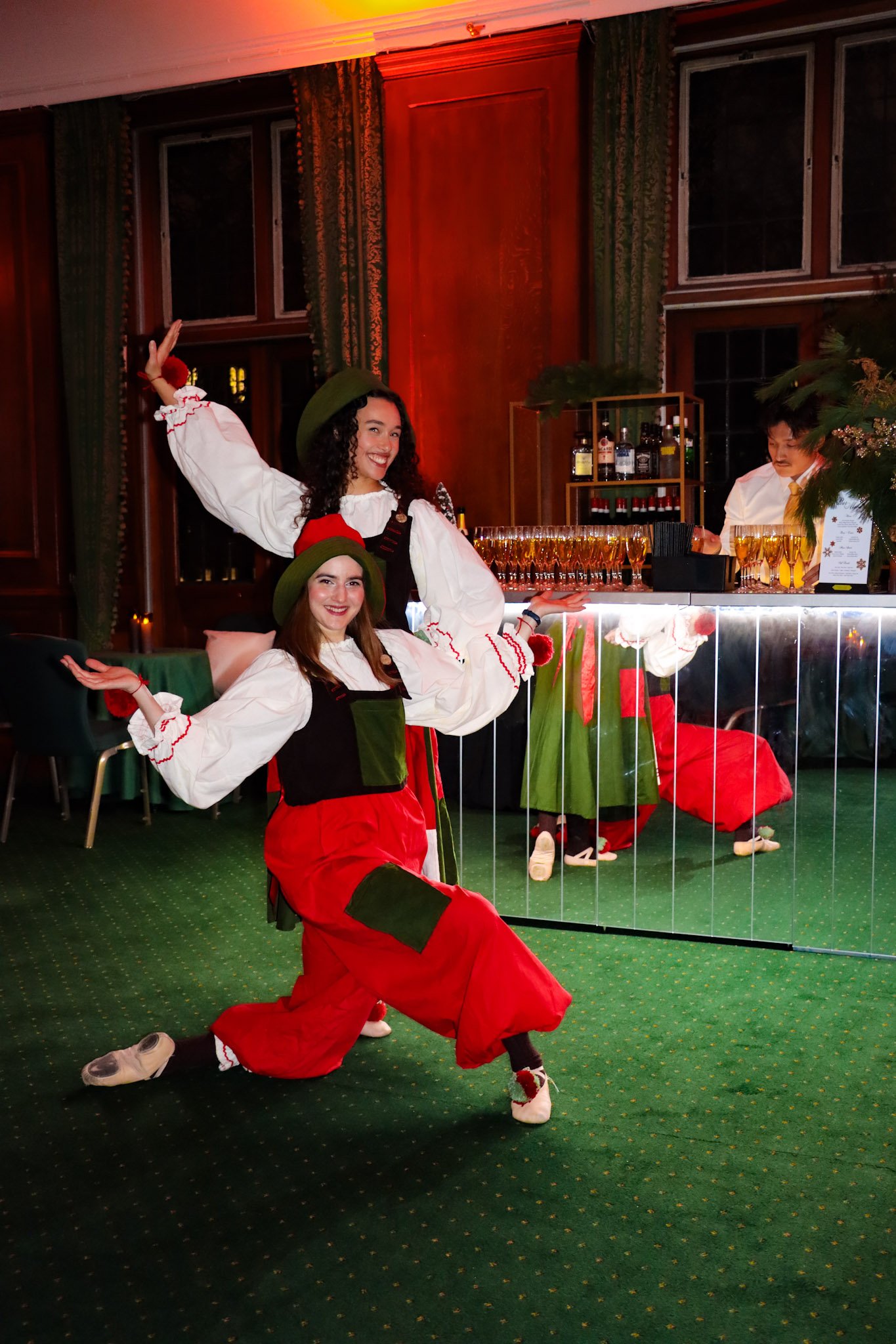 Two women in festive costumes performing a dance in a decorated room. One is kneeling on the green carpeted floor with arms outstretched, the other standing behind and smiling, both wearing red, green, and white outfits with wide-brimmed hats.