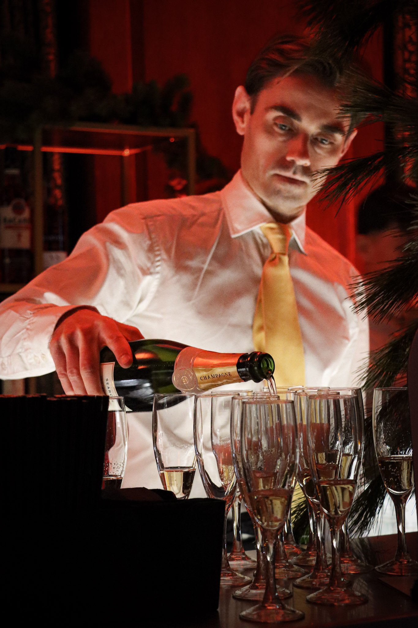 A man with dark hair, wearing a white shirt and yellow tie, pouring champagne into glasses on a table with multiple empty glasses and a black bucket, with red lighting and greenery in the background.