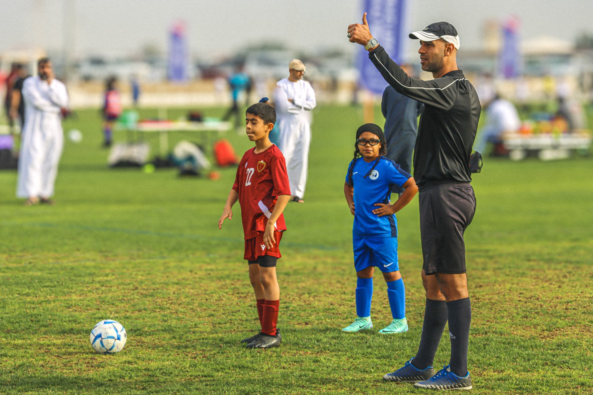A soccer coach giving instructions to young players on the field during daytime. Two children, a boy in a red uniform and a girl in a blue uniform, are standing with the coach near a soccer ball on a grass field. Multiple people are visible in the ba