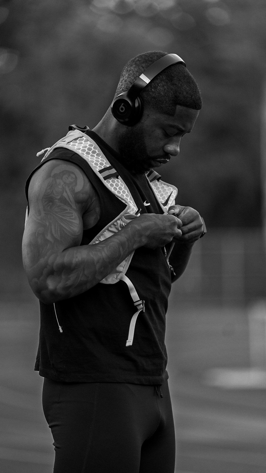 A man with a shaved hairstyle and tattoos on his arm, wearing headphones and adjusting his athletic vest in a gym or sports facility.