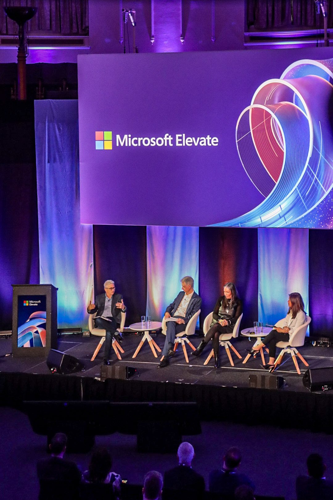 Panel discussion at Microsoft Elevate event with four speakers seated on stage, large screen displaying "Microsoft Elevate" logo with colorful graphic in background.