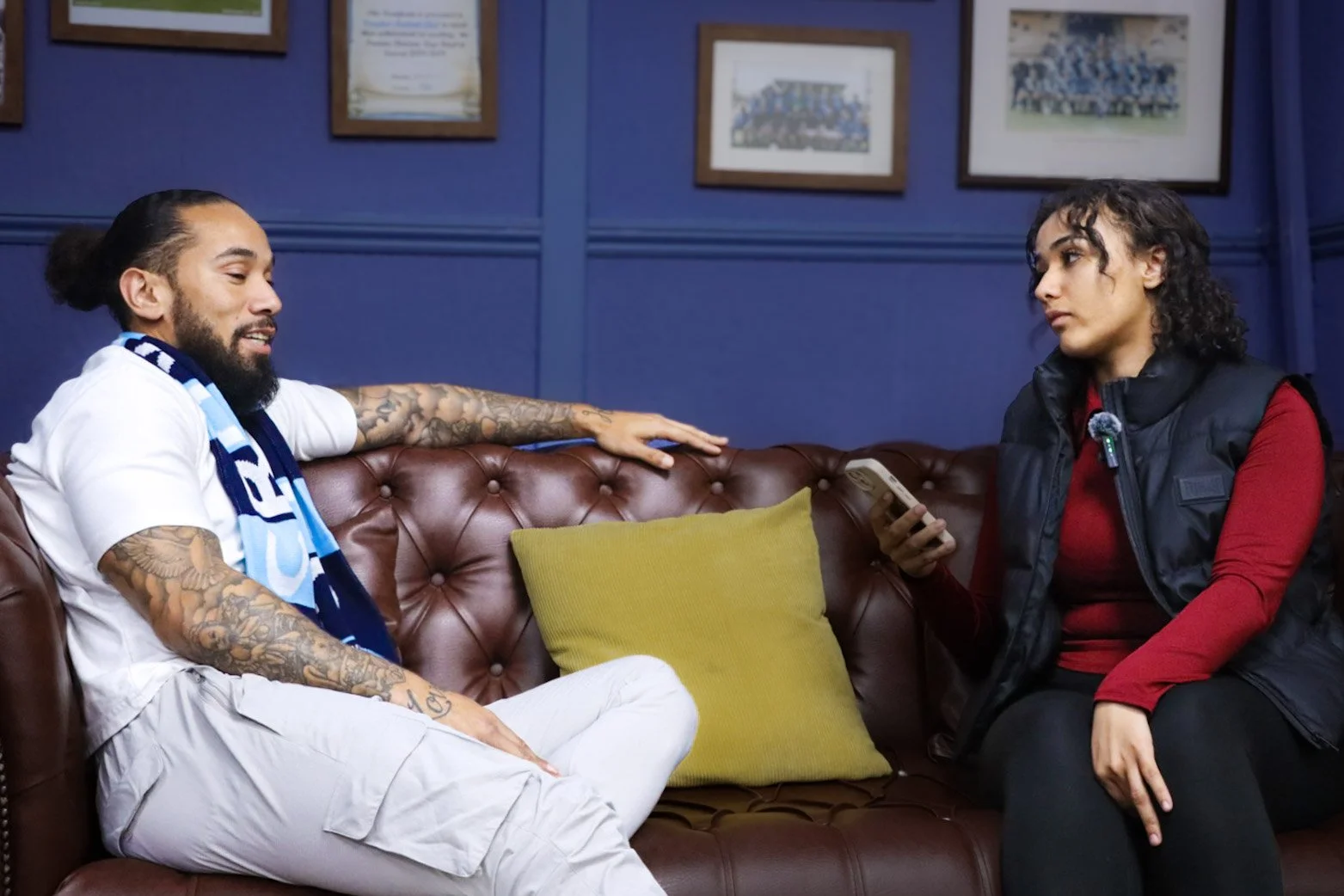 A man with tattoos, wearing a white shirt and a blue scarf, sitting on a brown leather couch, talking to a woman with curly hair, wearing a red top and black vest, holding a phone. They are in a room with blue walls and framed pictures.