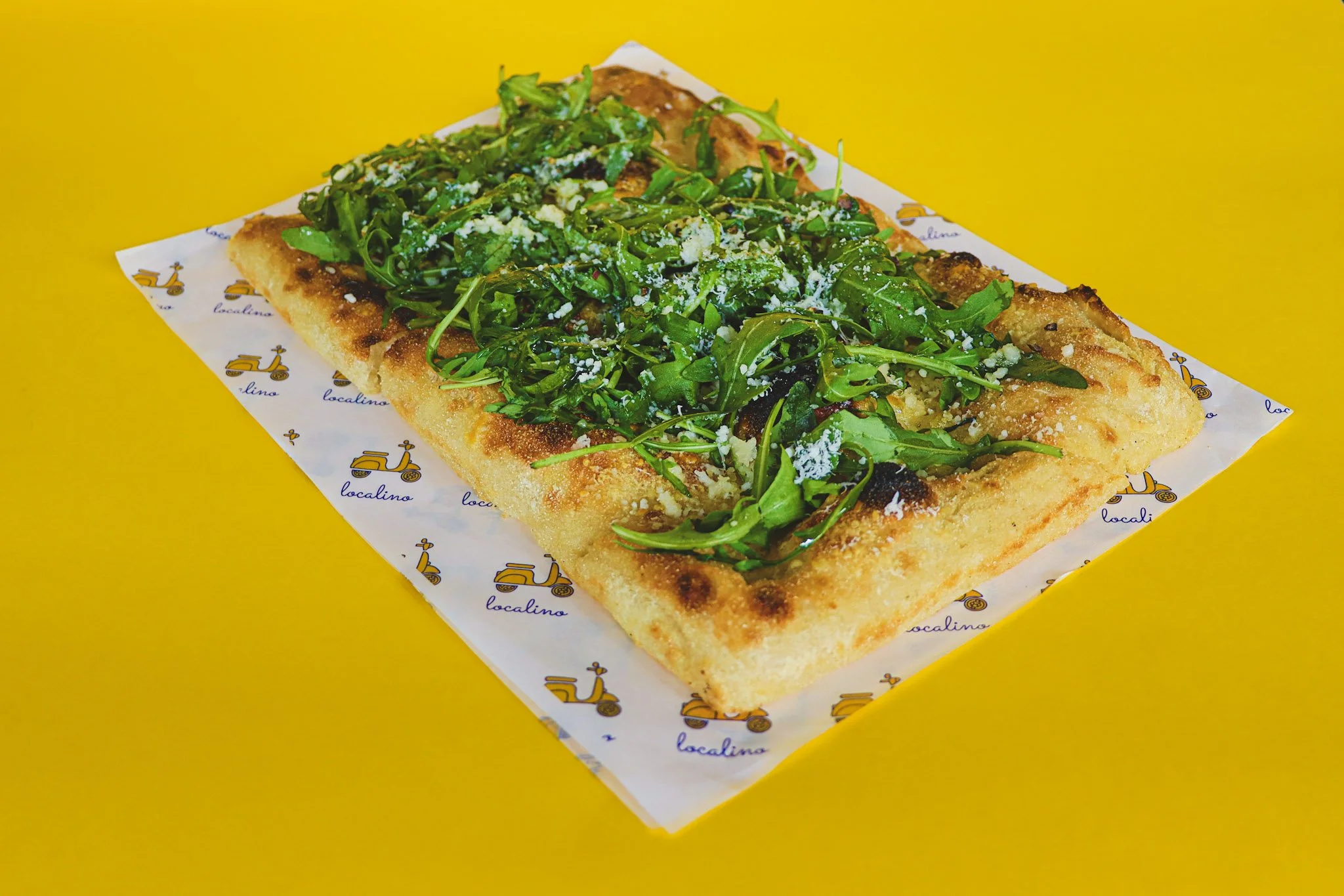 A rectangular pizza with a thick golden crust topped with fresh green arugula and grated cheese, placed on a white paper liner with a yellow background.