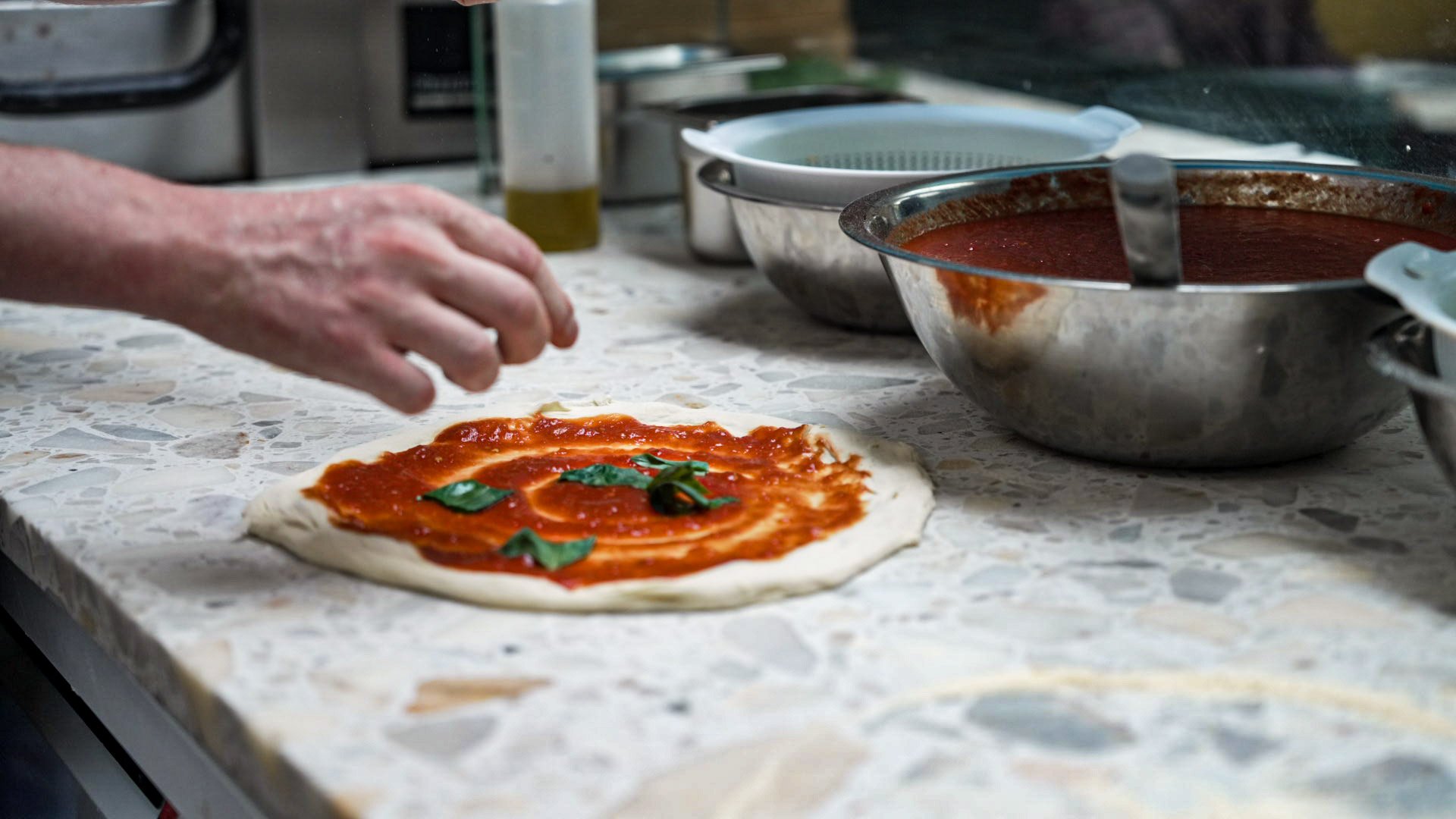 Person adding fresh basil leaves to a pizza with tomato sauce and dough on a marble countertop