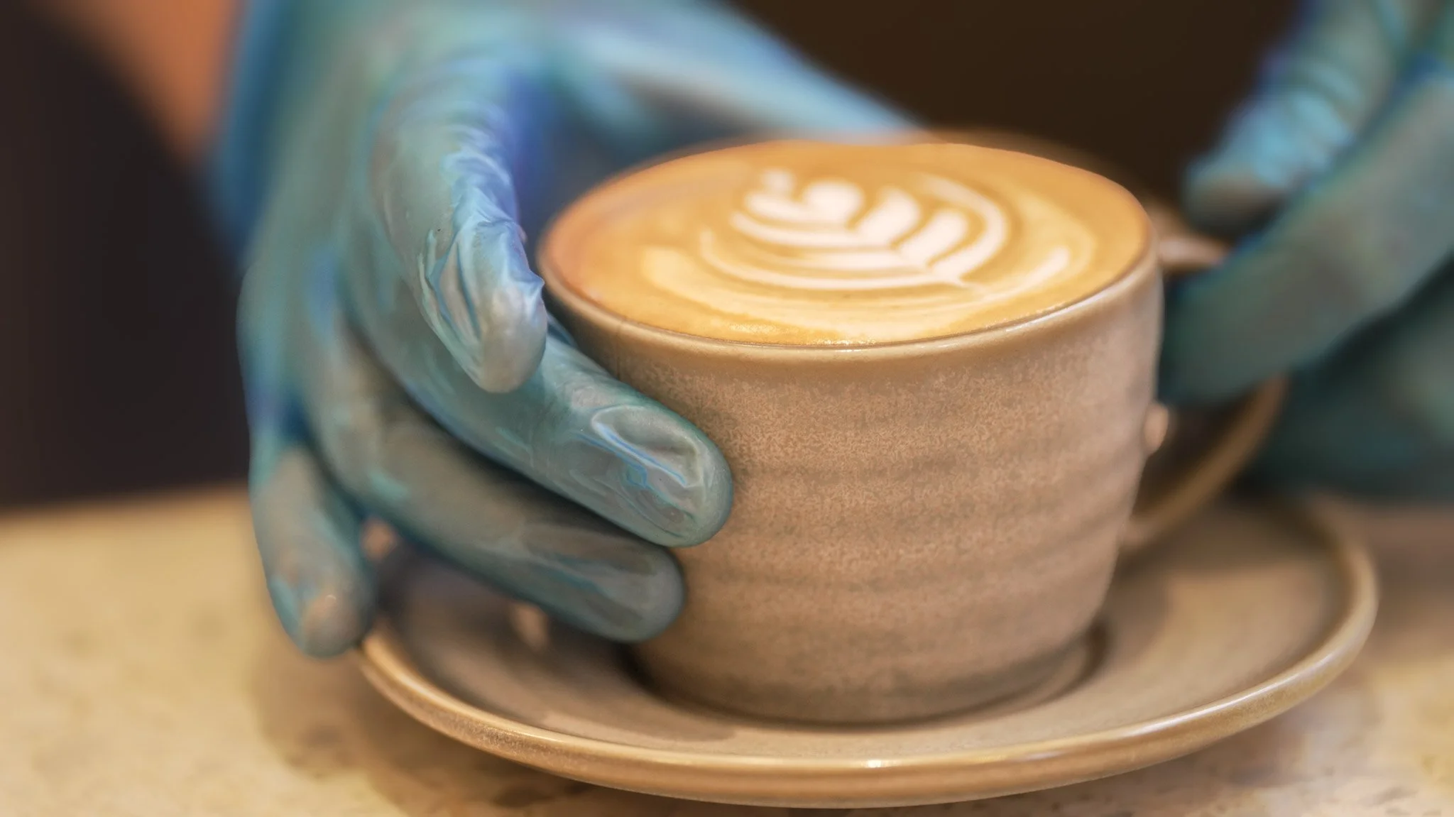 A person's hand wearing a blue glove holding a small ceramic cup of coffee with latte art on top, placed on a matching saucer.