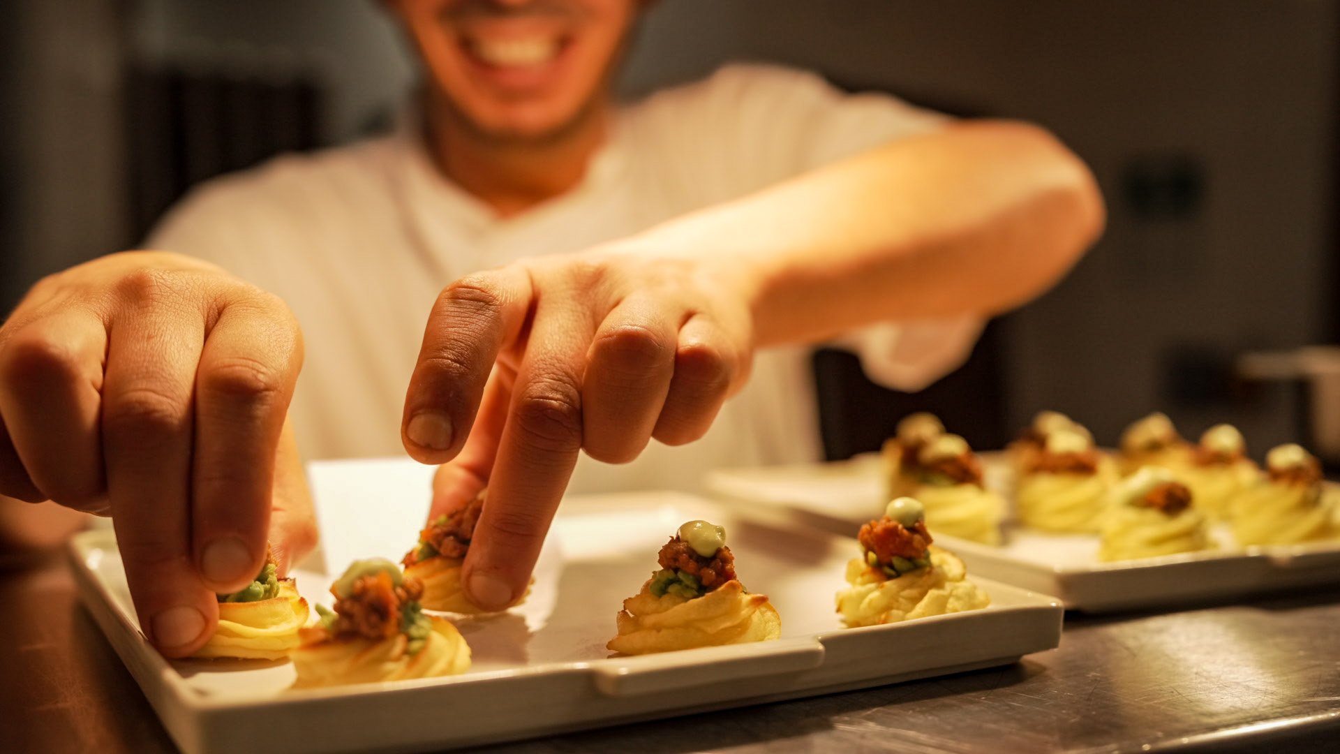 Close-up of a person's hands decorating canapé appetizers on a white serving platter, with a smiling person in the background and multiple similar appetizers on the platter.