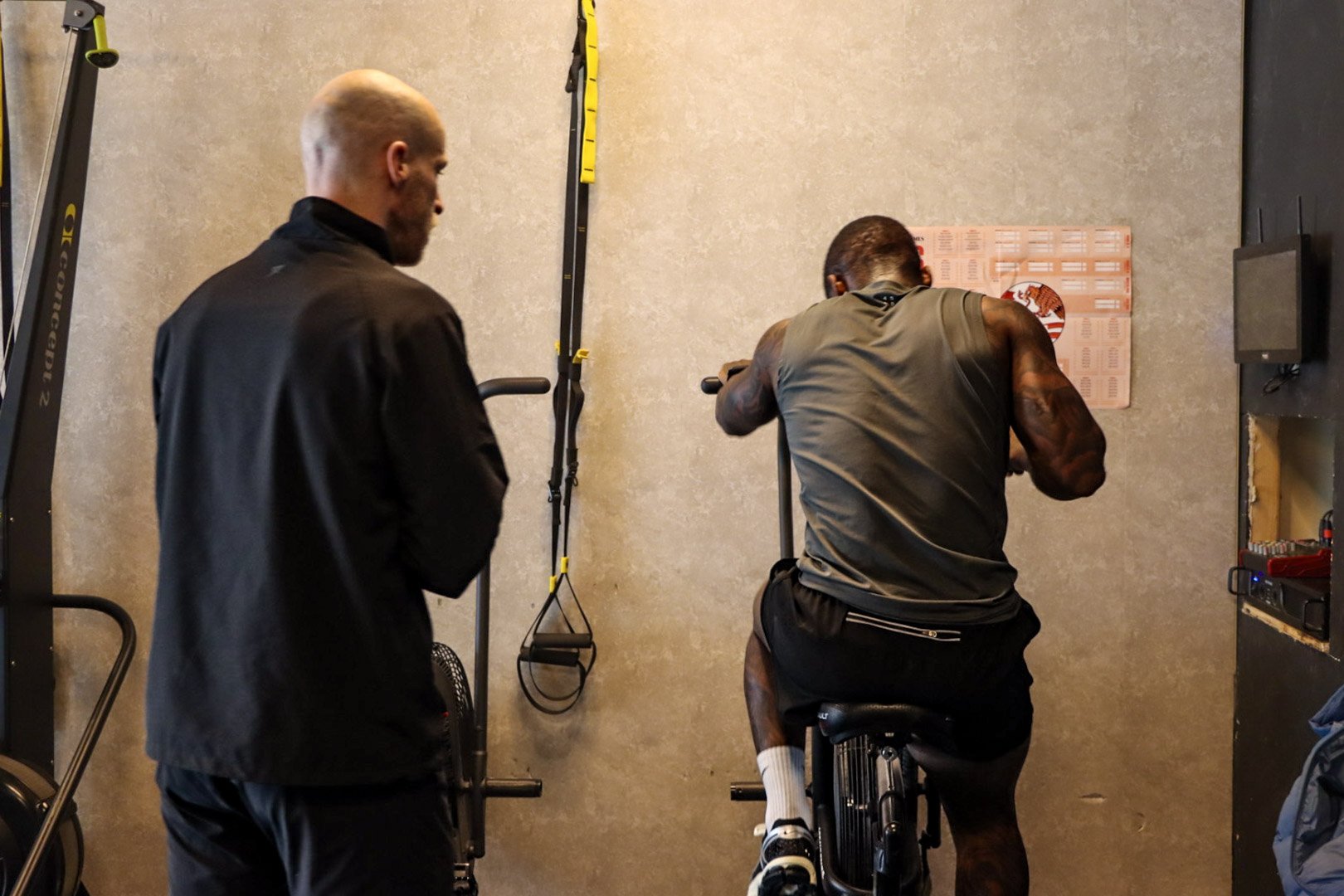 Two men in a gym, one sitting on a stationary bike and stretching, the other standing nearby watching.