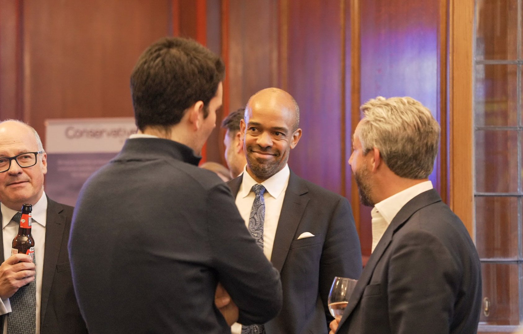 A group of middle-aged men in suits and ties are talking and smiling at a social or professional event in a wood-paneled room. One man holds a beer, another a glass of wine, and a person in back is slightly blurred.