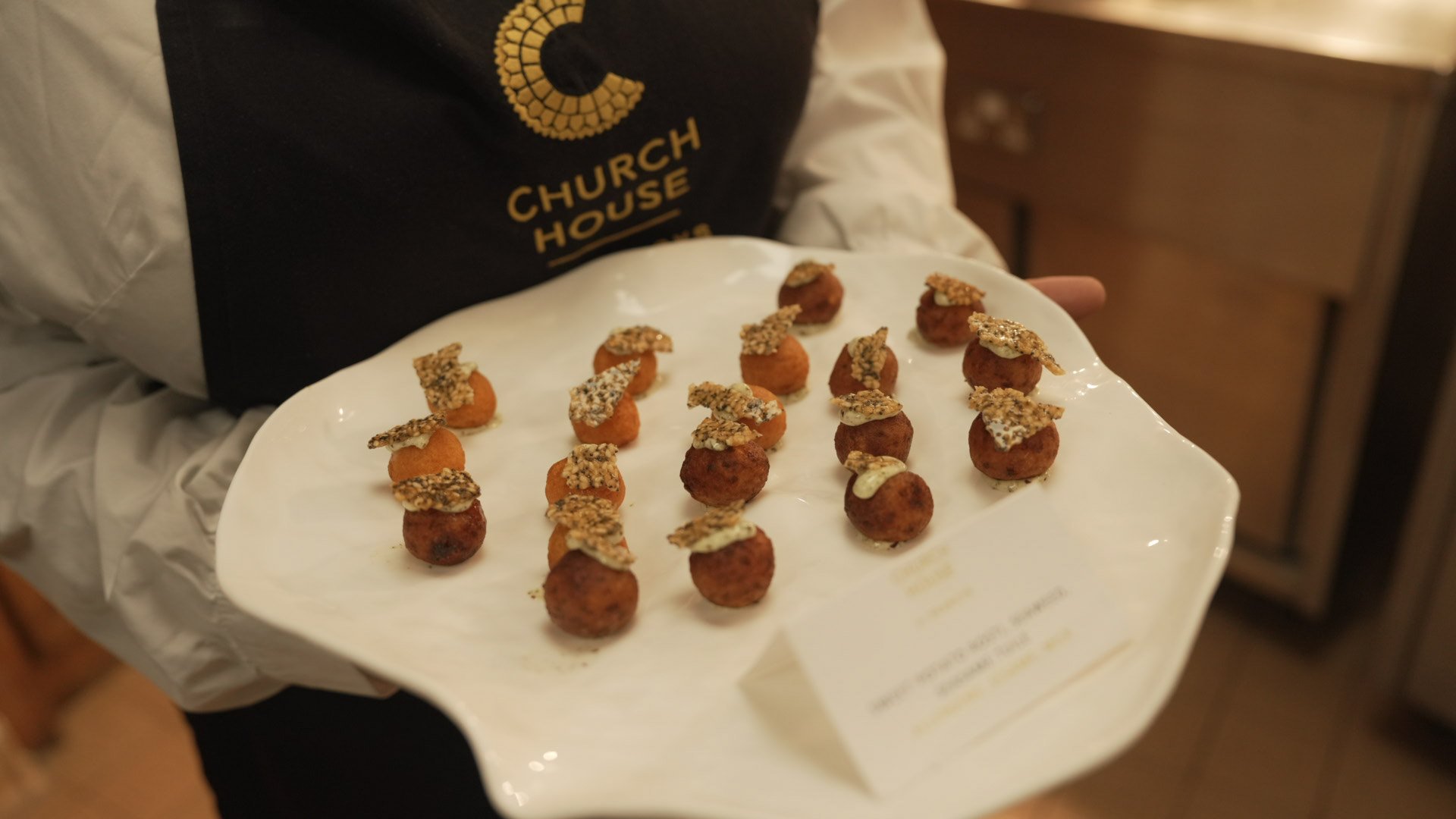 Plate of bite-sized appetizers consisting of round balls topped with a crispy garnish, served on a white plate held by a person wearing a black apron with gold lettering and a white shirt.