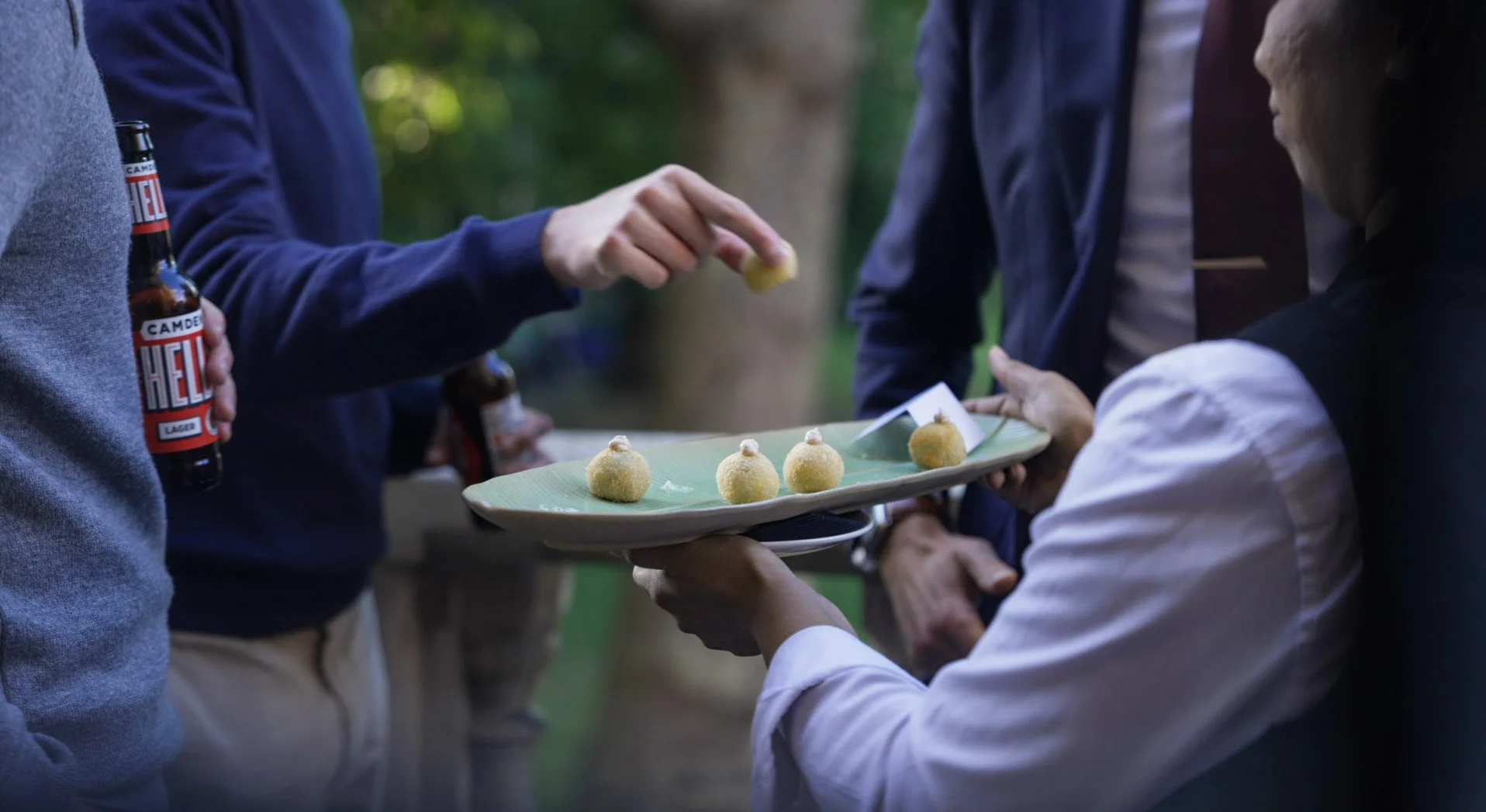 People at an outdoor gathering sharing a tray of round, breaded appetizers, with one person handing over a piece to another, in a park or garden setting.