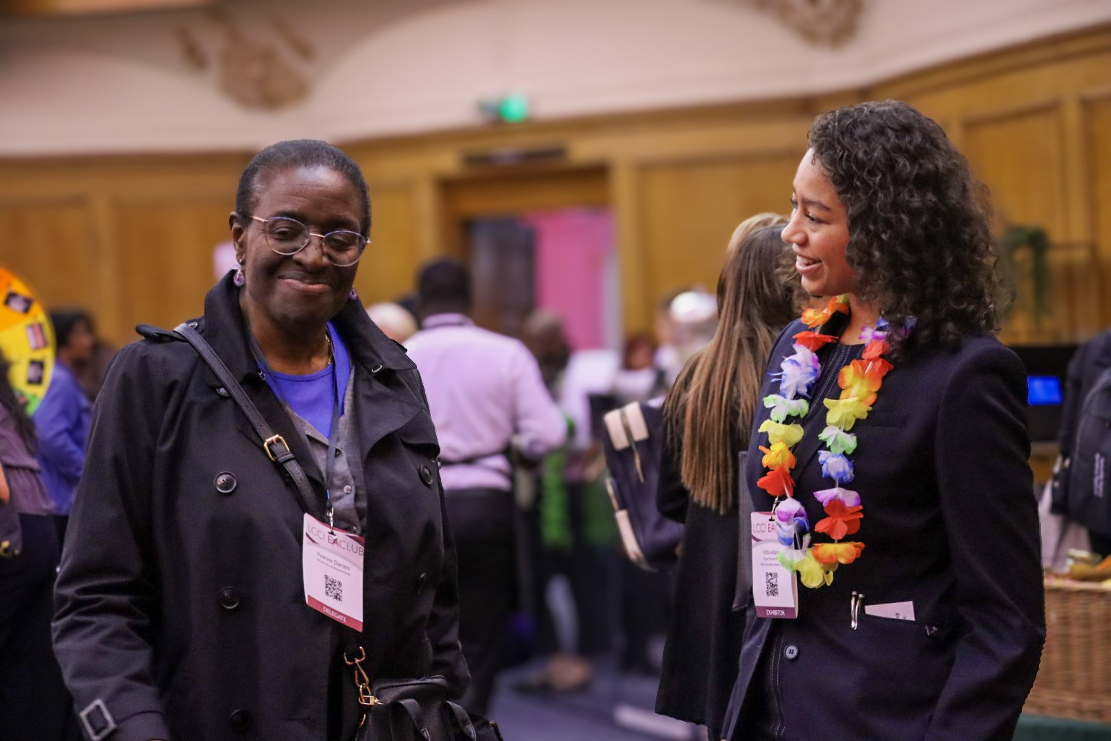 Two women smiling and talking at a conference. The woman on the right is wearing a colorful lei, and both are wearing conference badges.