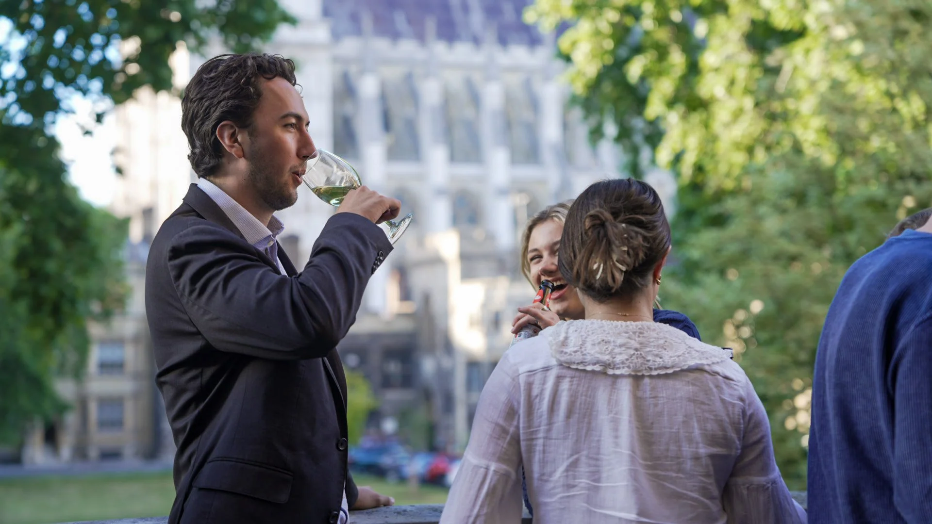 A man in a dark suit and light shirt is drinking white wine outdoors during daytime, with city buildings and greenery in the background, while talking to two women with their back to the camera.
