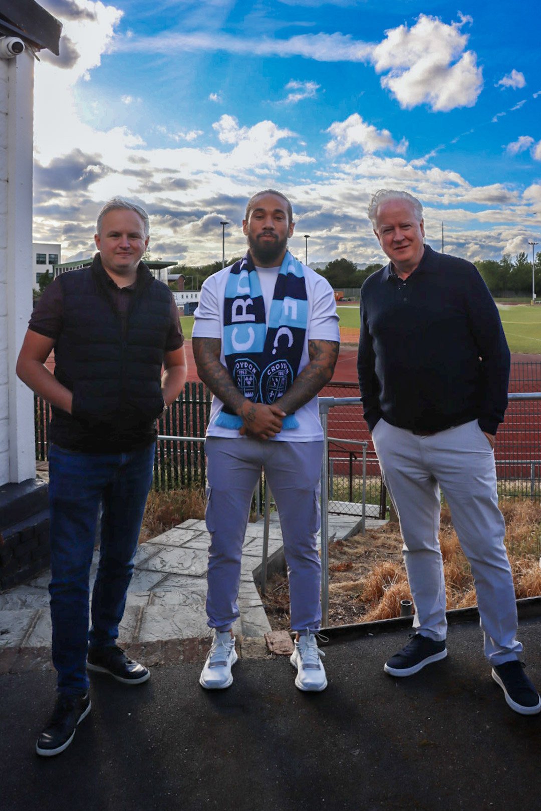 Three men standing outdoors on a track field with a fence, greenery, and a partly cloudy sky in the background. The man in the center is wearing a white shirt, light gray pants, and a blue and white scarf.