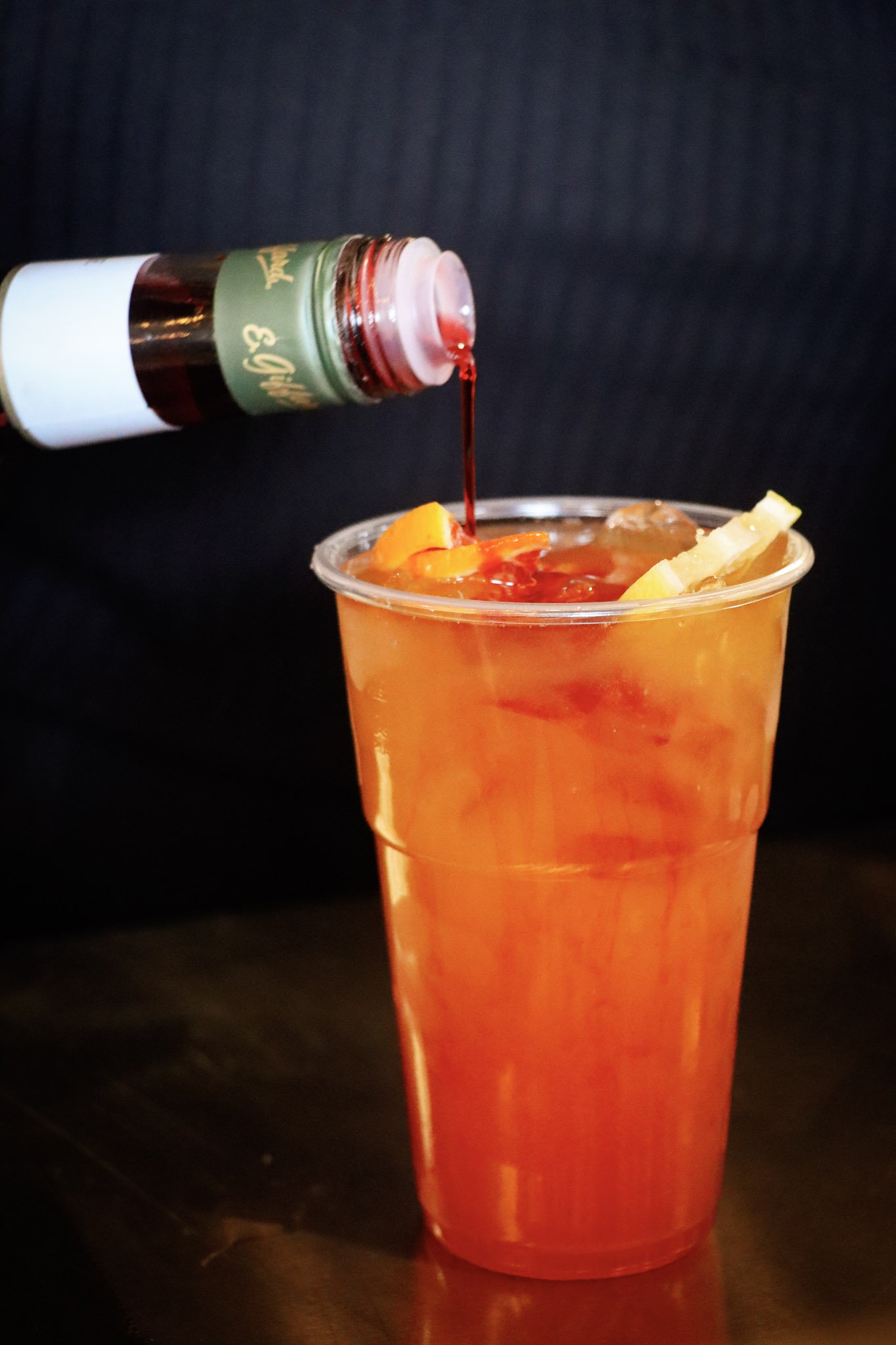 A cup filled with orange-colored drink and ice, with pineapple pieces on top, and a bottle pouring red syrup into it.