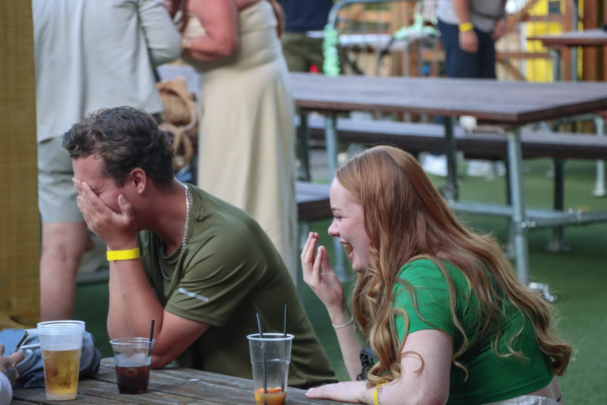 Two young adults, a man and a woman, are sitting at a picnic table outdoors, laughing and covering their faces, with drinks in front of them. They seem to be enjoying a fun moment together.