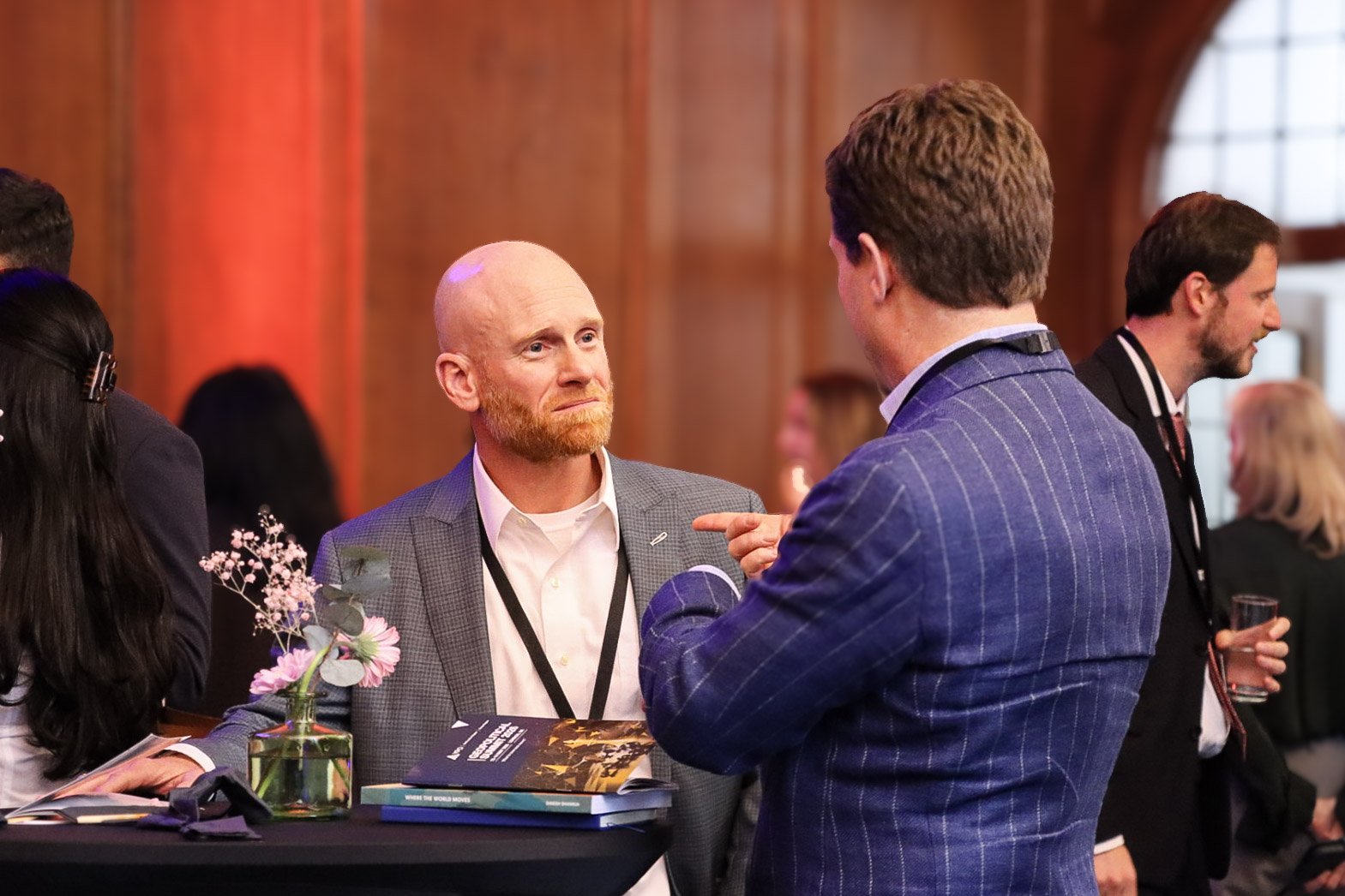 Two men are engaged in a conversation at a professional event or conference. One man is bald with a beard, wearing a gray checkered blazer and white shirt. The other man has short dark hair, wearing a blue striped blazer. They are standing at a table