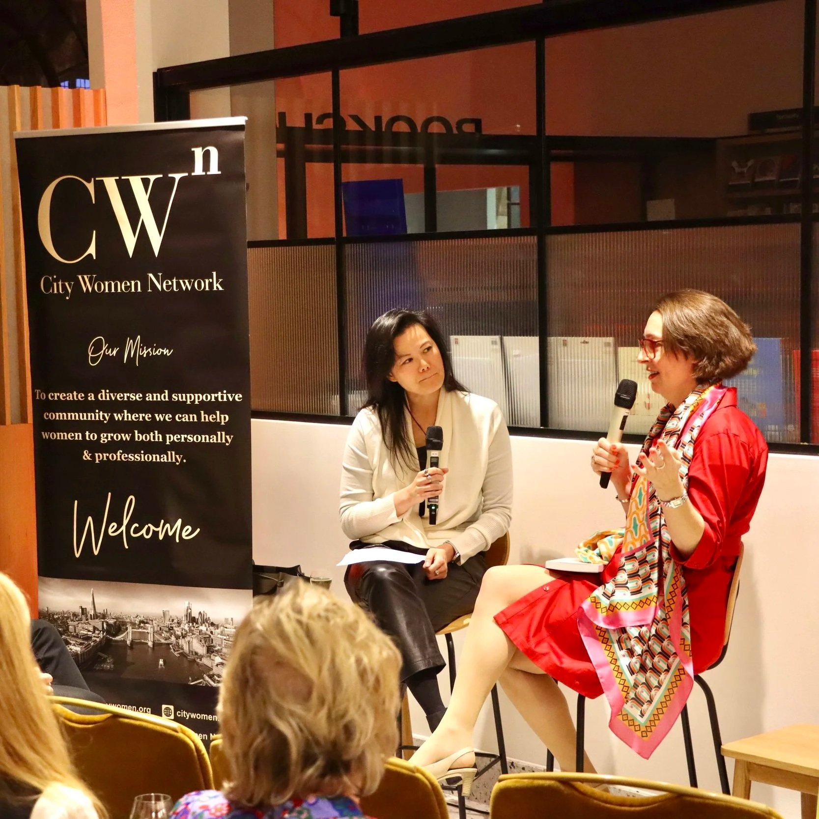 Two women are seated and speaking into microphones during a panel discussion at an event organized by City Women Network, with a black banner displaying the organization's logo and mission statement in the background.