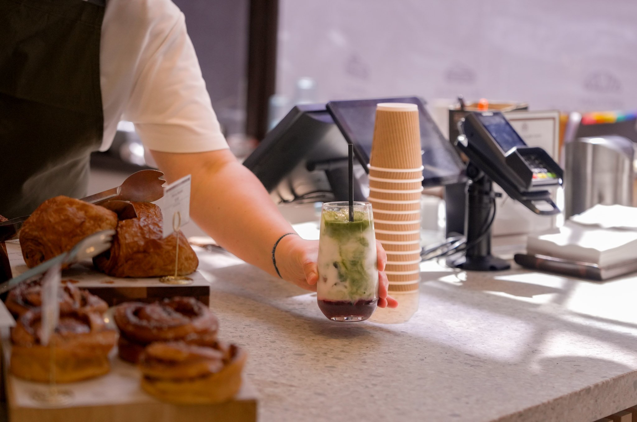 A person holding a layered drink with mint, lime, and other ingredients in a tall glass with a straw at a coffee shop or cafe counter. There are stacks of disposable cups, point-of-sale screens, and baked goods on display nearby.