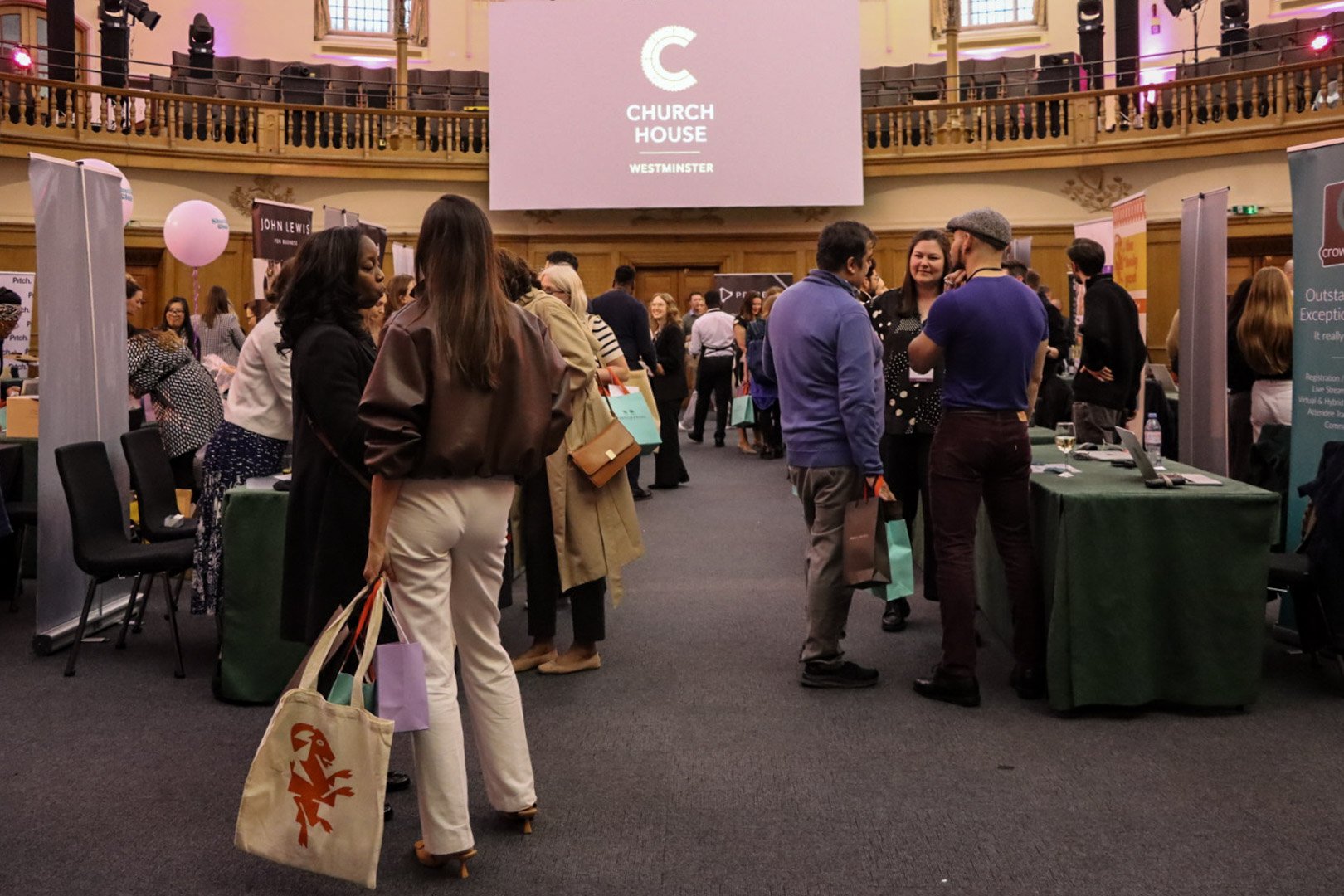 People attending an event in a historic church hall with booths and a large screen displaying 'Church House Westminster'
