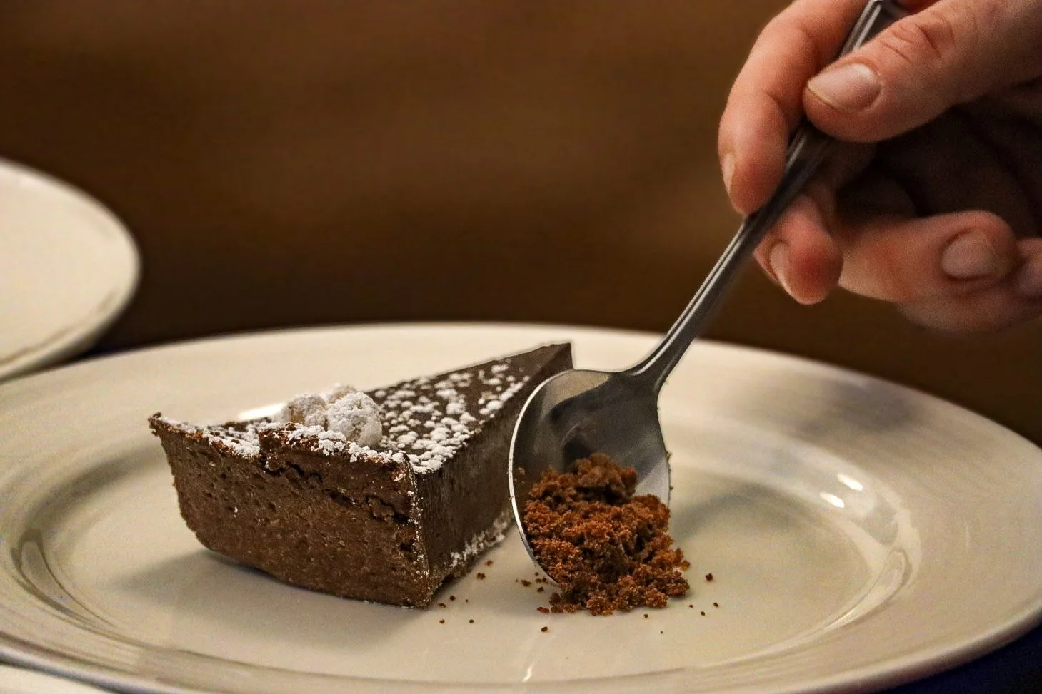 A slice of chocolate cake with powdered sugar and a dollop of whipped cream on top on a white plate. A hand is holding a spoon filled with crumbled chocolate cookie or cake crumbs, about to place them on the plate.