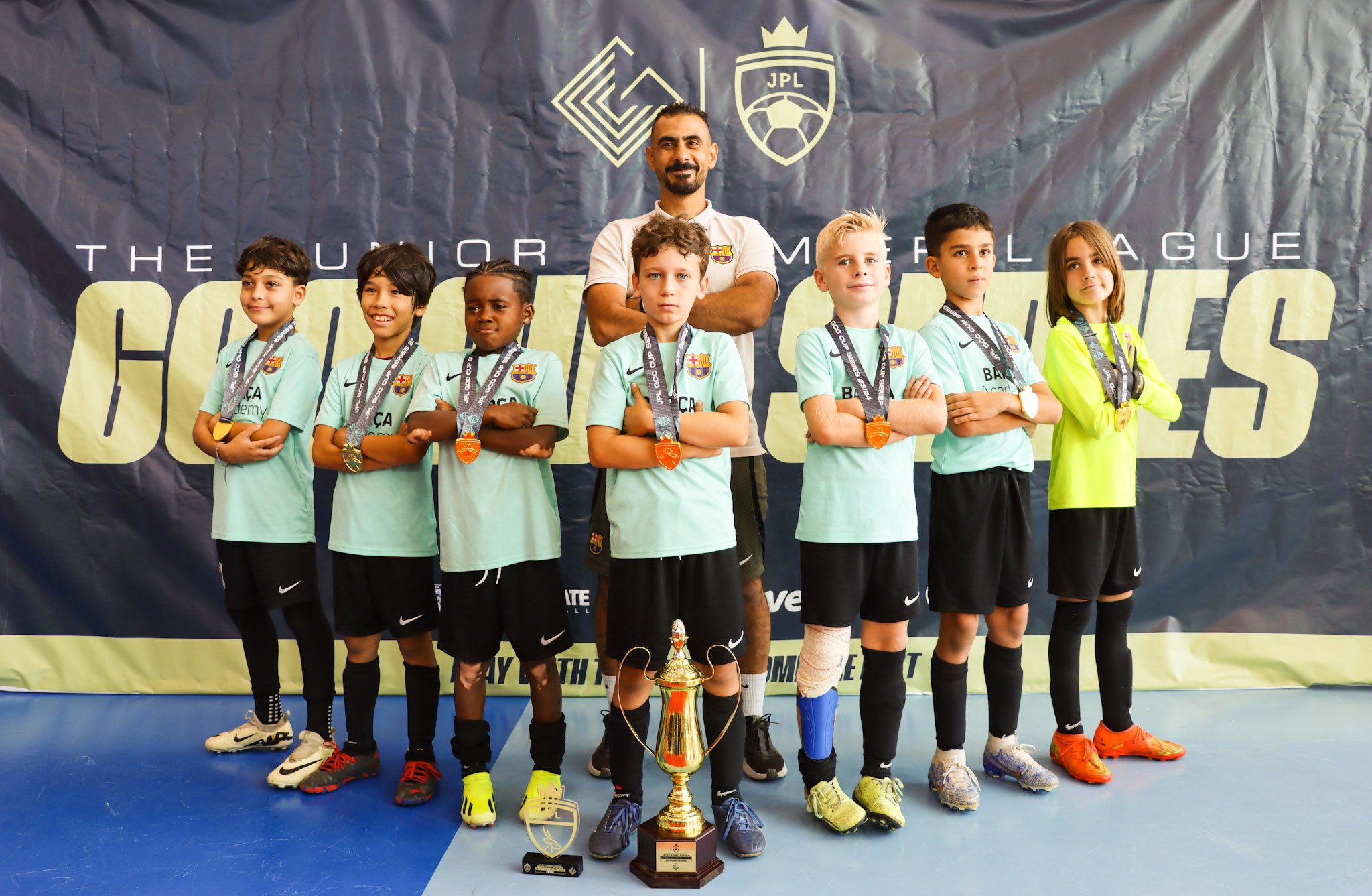 A group of young children in soccer uniforms standing in front of a banner, with a coach behind them. They are all wearing medals around their necks and are standing with crossed arms. There is a trophy placed in front of them on the floor.