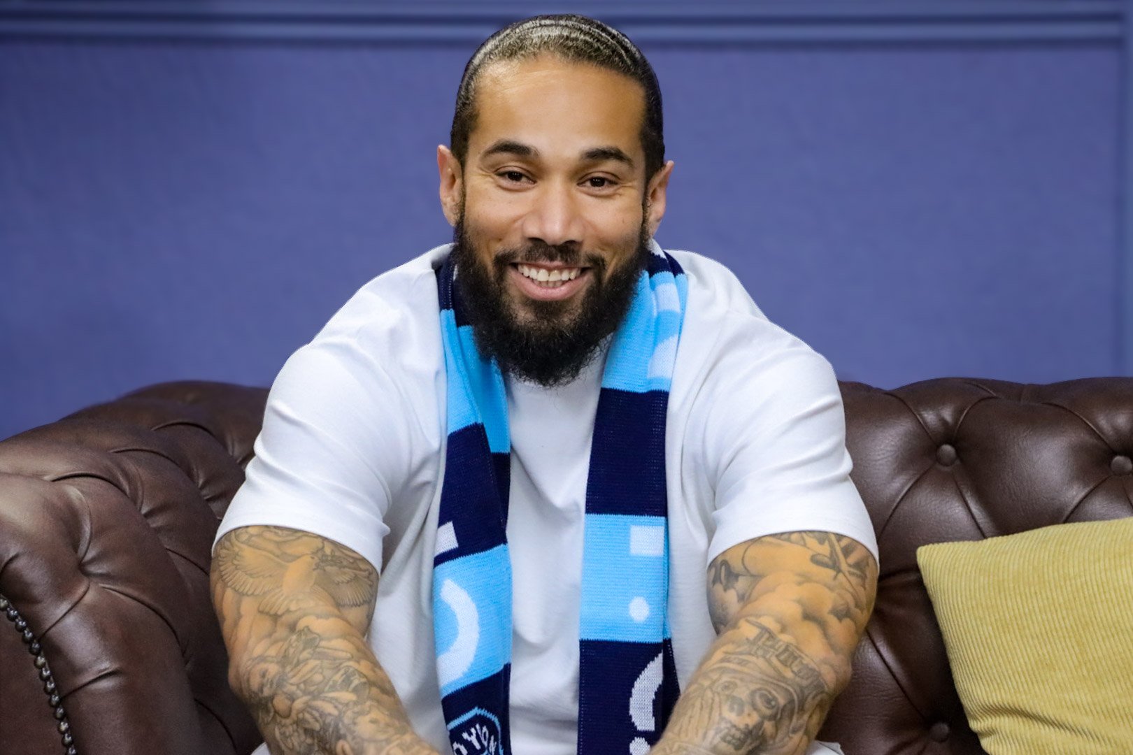 A smiling man with a beard and tattoos on his arms sitting on a brown leather couch, wearing a white t-shirt and a blue scarf with light blue and dark blue patterns, in front of a blue background.