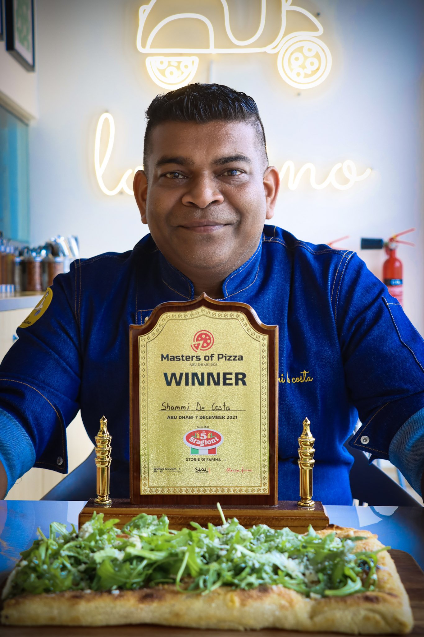 A man in a blue shirt holding a pizza with arugula and other greens. Behind him, a neon sign and a certificate or award is displayed.