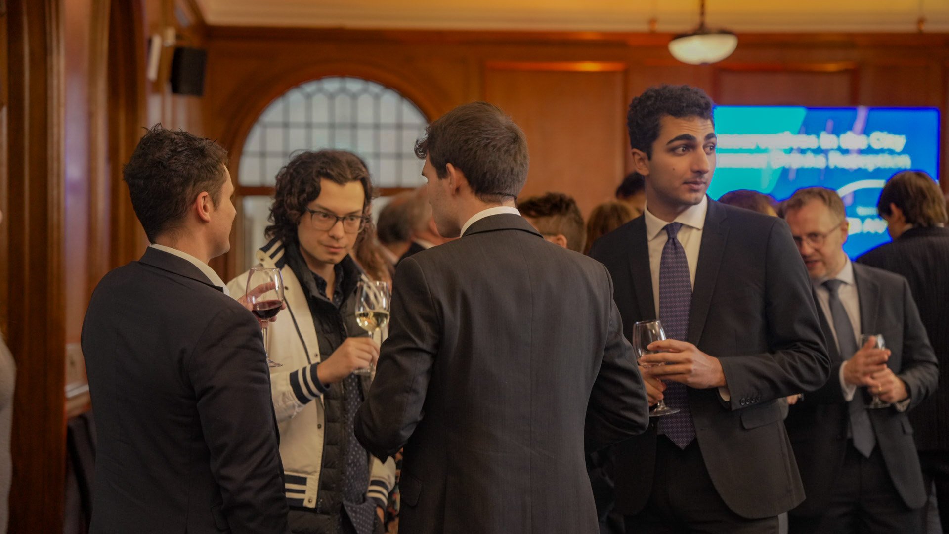 Business professionals at a networking event, wearing suits, holding wine glasses, and engaging in conversation in a wood-paneled room.