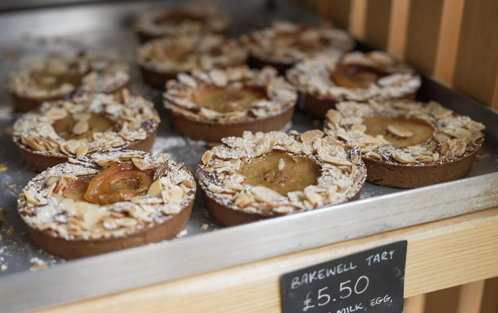 Several almond tart pastries with a golden filling and sliced almonds on top, displayed on a metal tray at a bakery.