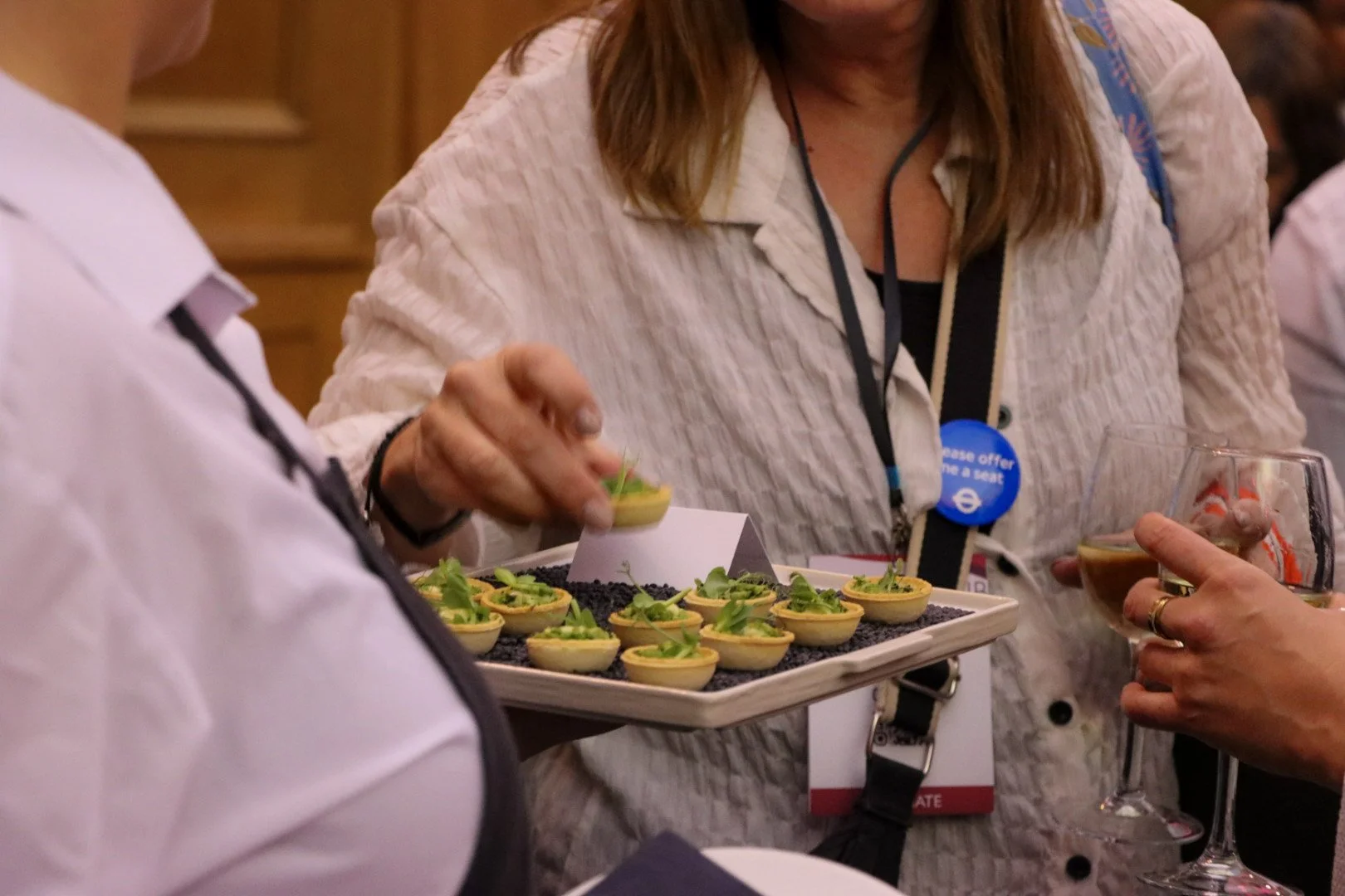 People at a social event interacting, with a tray of small appetizers or cups of food and drinks in hand.