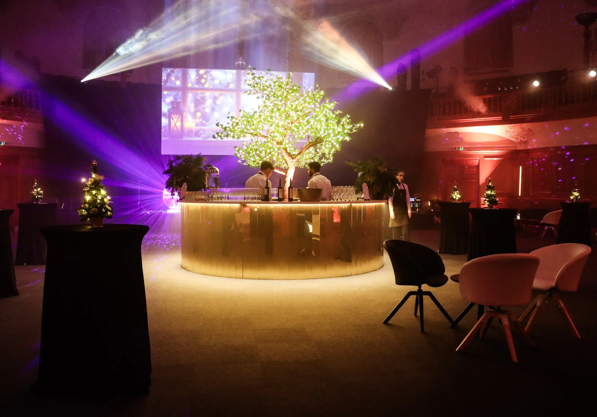 An indoor event space decorated for a festive occasion with illuminated trees, small decorated Christmas trees, and colorful lighting effects.