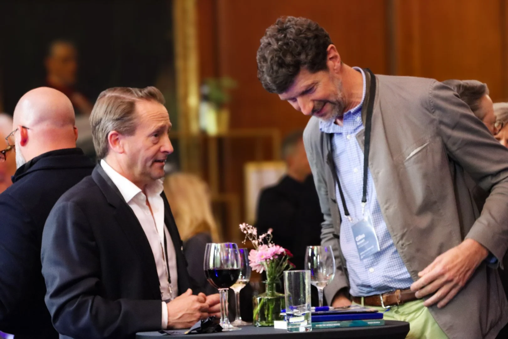 Two men are engaged in conversation at a dining event, standing by a table with wine glasses and a small floral arrangement. Other guests are visible in the background.