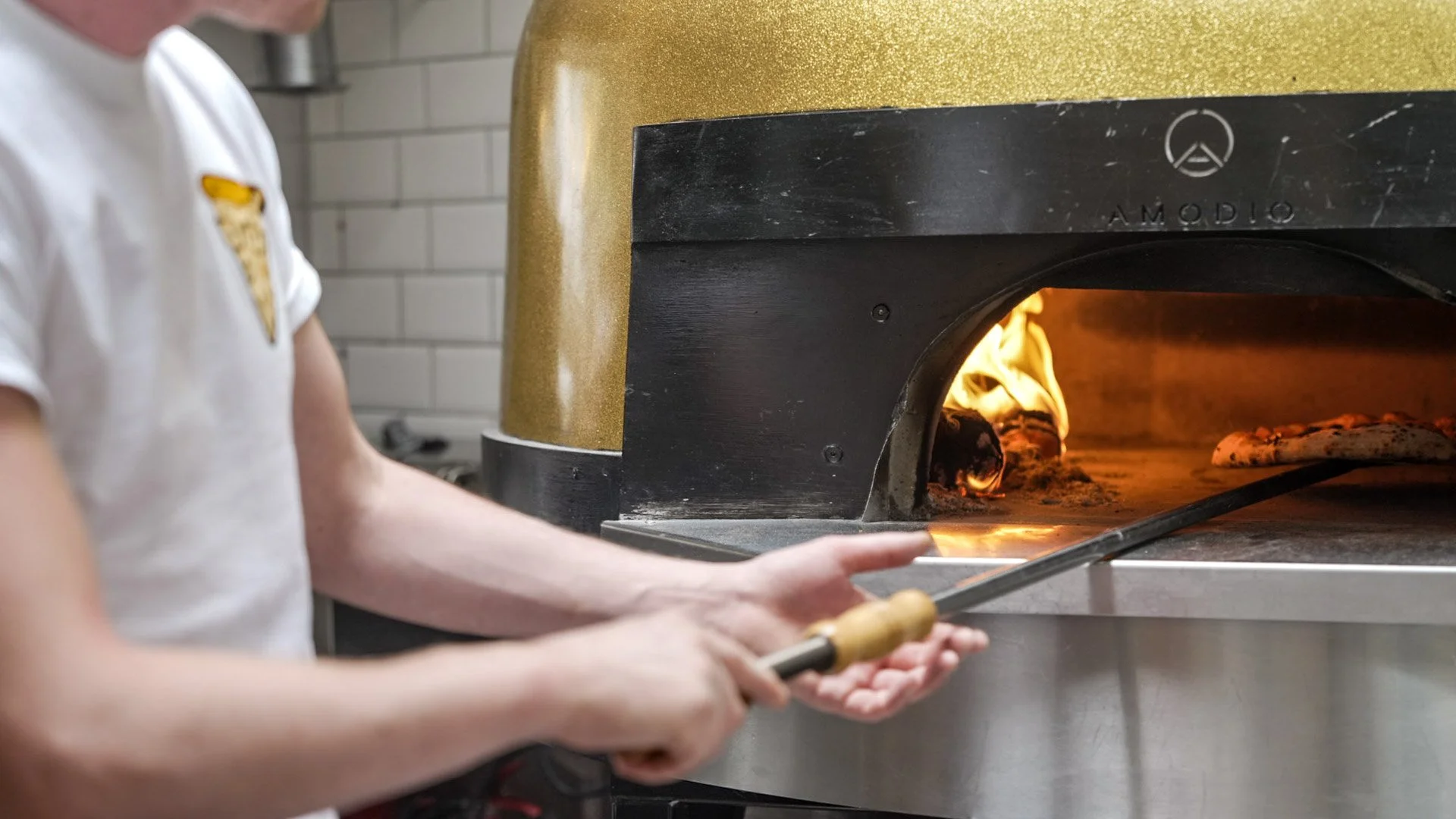 Person placing a pizza inside a wood-fired oven with flames visible inside.