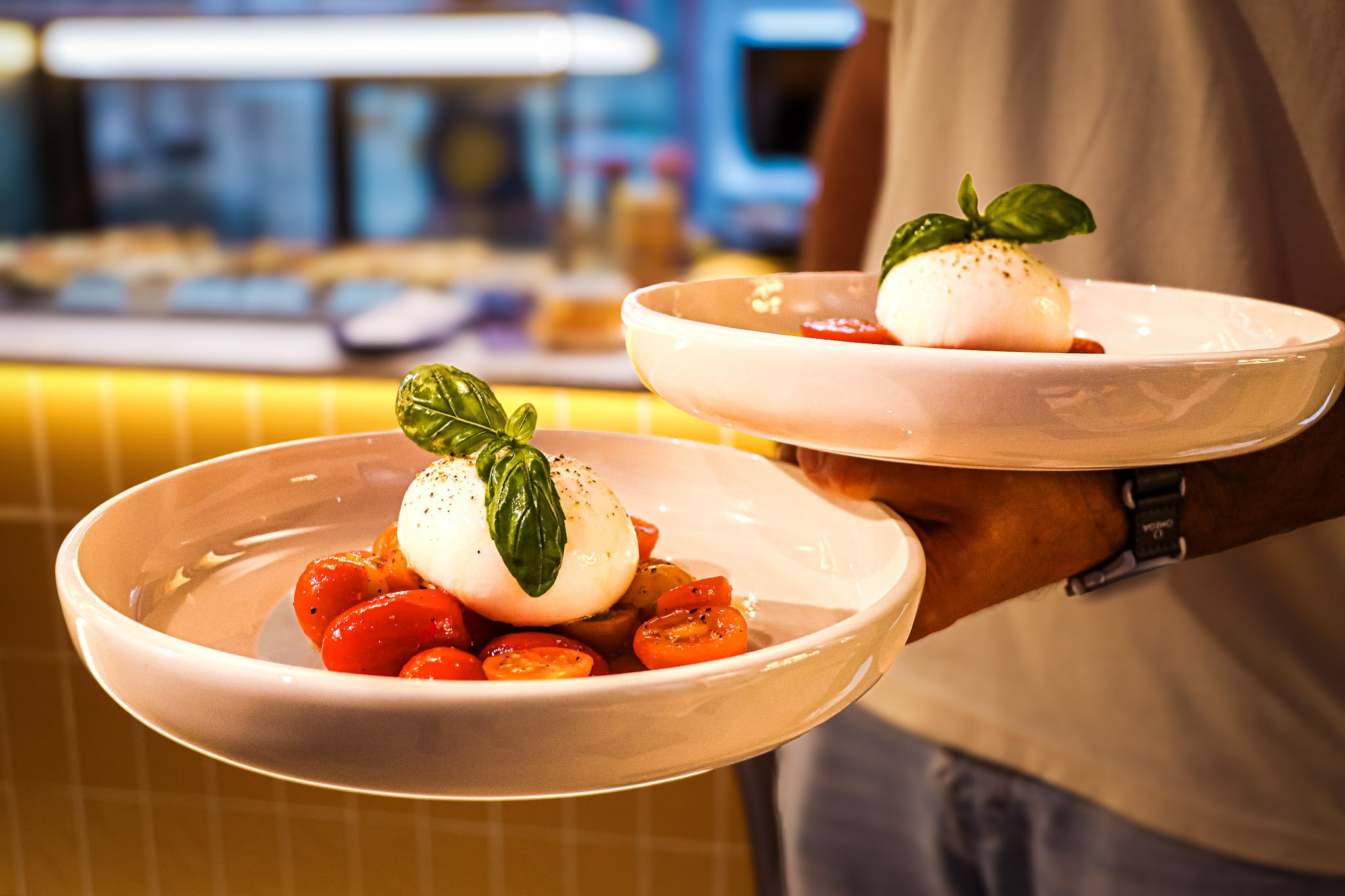 Two white bowls with cherry tomatoes, mozzarella balls, and basil leaves. One bowl is slightly elevated on a tray. Blurry kitchen background.