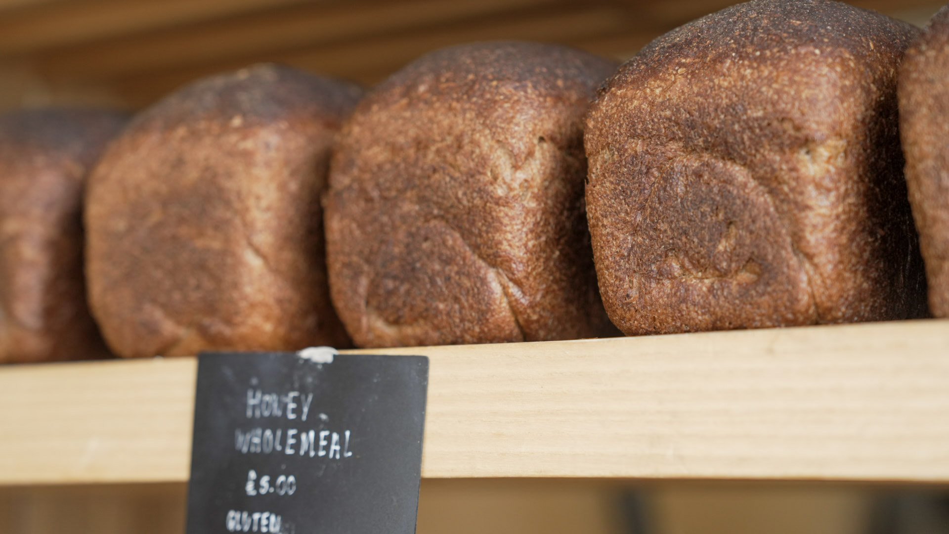Close-up of several brown bread rolls or loaves on a wooden shelf in a bakery, with a black chalkboard sign showing prices and information.