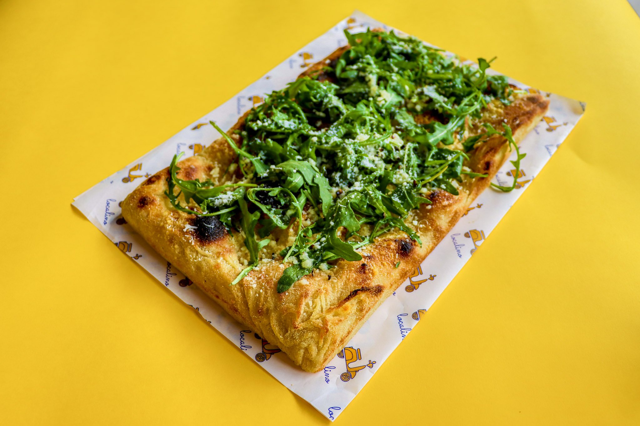 A rectangular pizza topped with arugula and grated cheese on a piece of branded paper on a yellow surface.