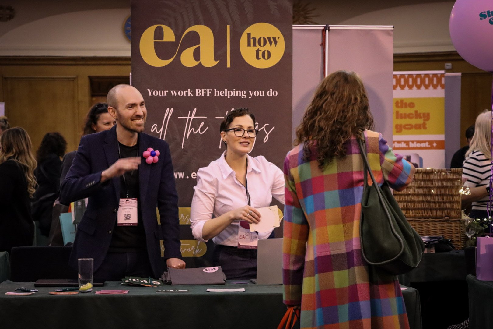 At a conference booth, a smiling man and woman are engaging with a visitor. The booth has banners with marketing material, including one that reads 'ea | how to' and 'Your work BFF helping you do all the things.' There are baskets, a laptop, and vari