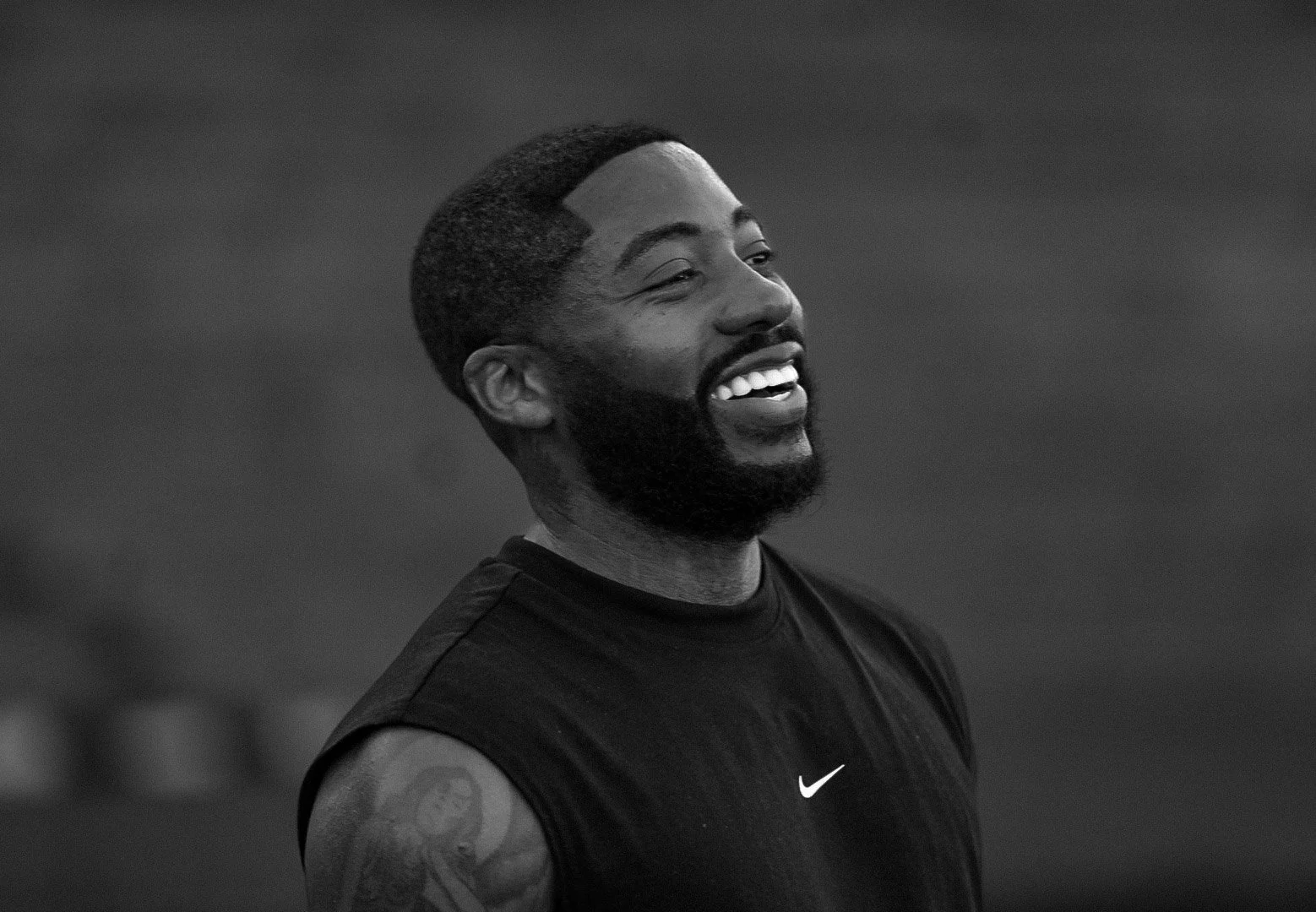 A smiling African-American man with a beard, wearing a sleeveless athletic shirt with the Nike logo, in a black and white photo.